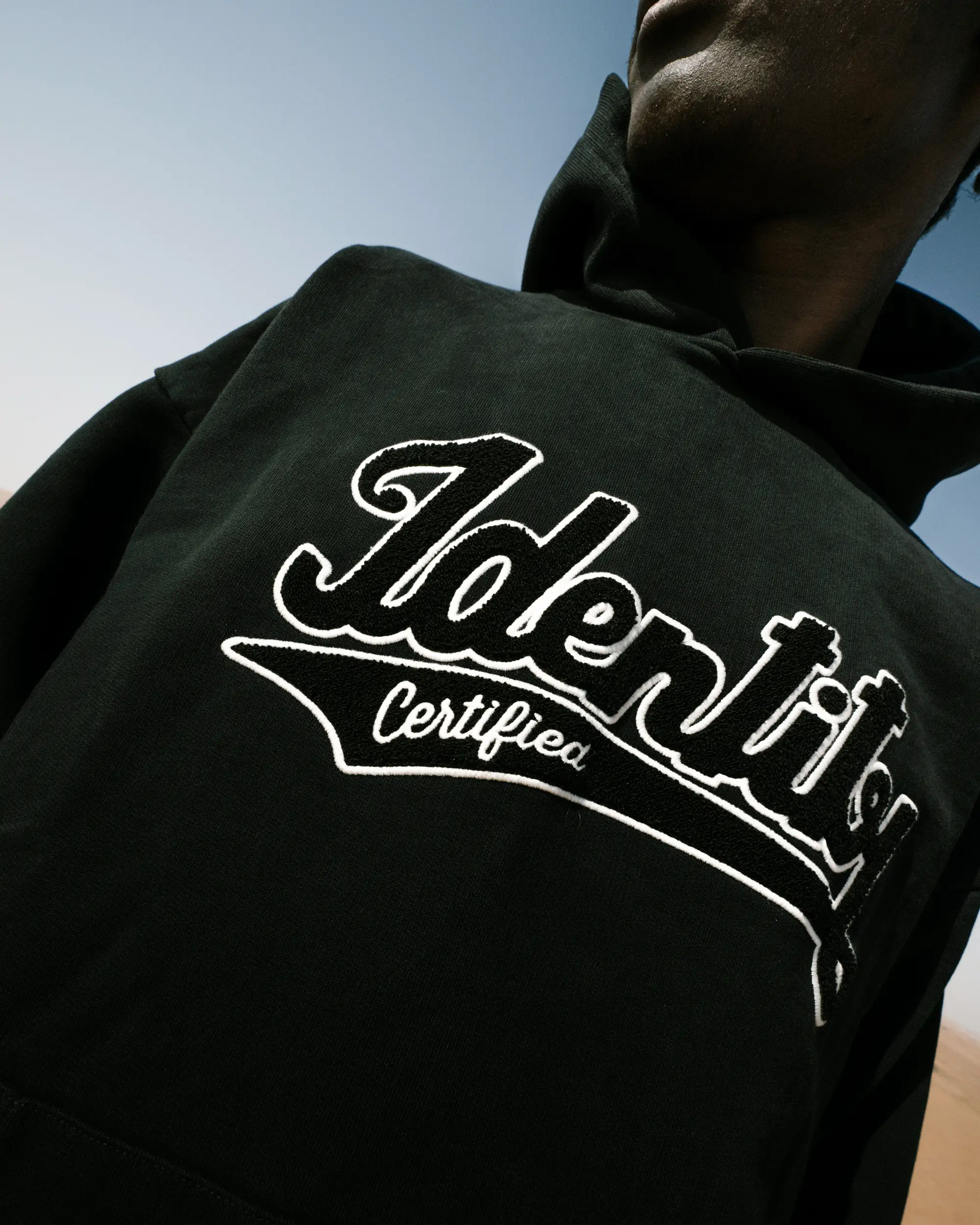 Nahaufnahme des weißen Identity-Schriftzugs auf Ibo Ouro-Bodis schwarzem Hoodie vor blauem Himmel.