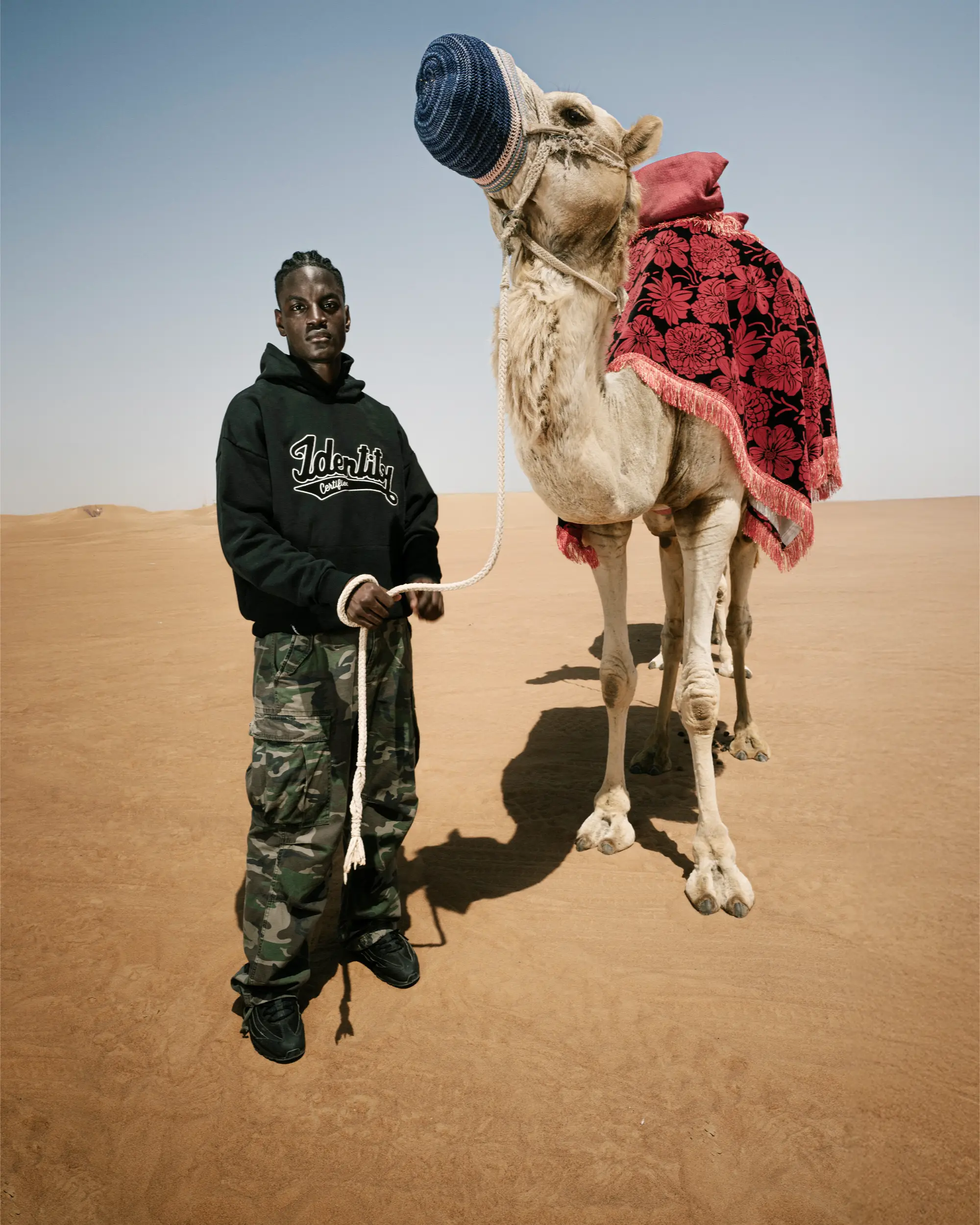 Ibo Ouro-Bodi im schwarzen Certified-Identity-Hoodie neben einem Kamel mit roter Satteldecke in der Wüste.