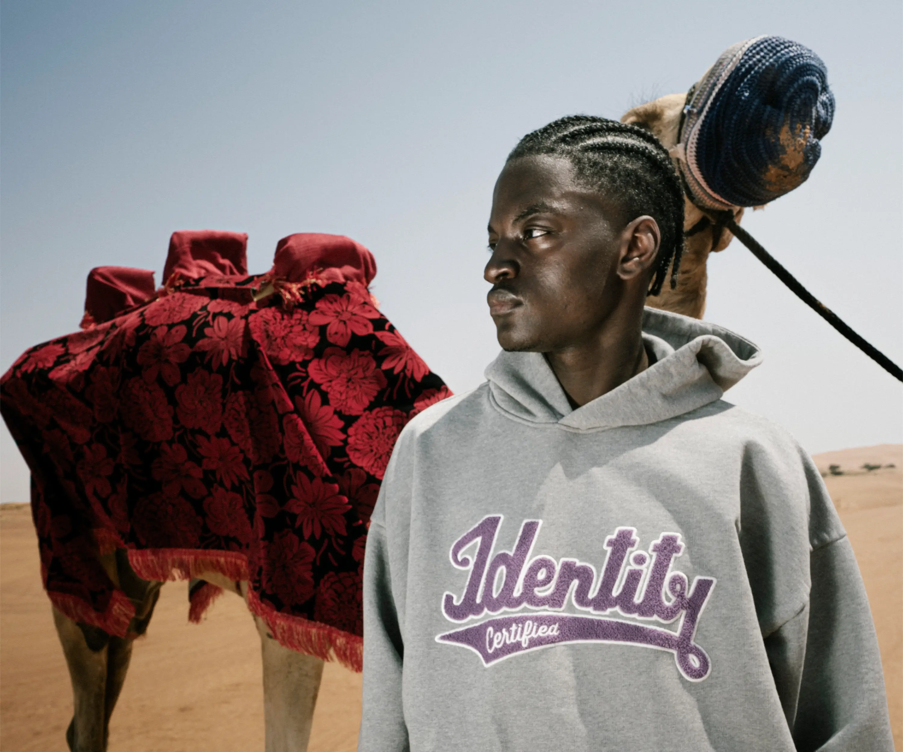 Ibo Ouro-Bodi im grauen Hoodie mit lilafarbener Identity-Stickerei neben einem Kamel in der Wüste.