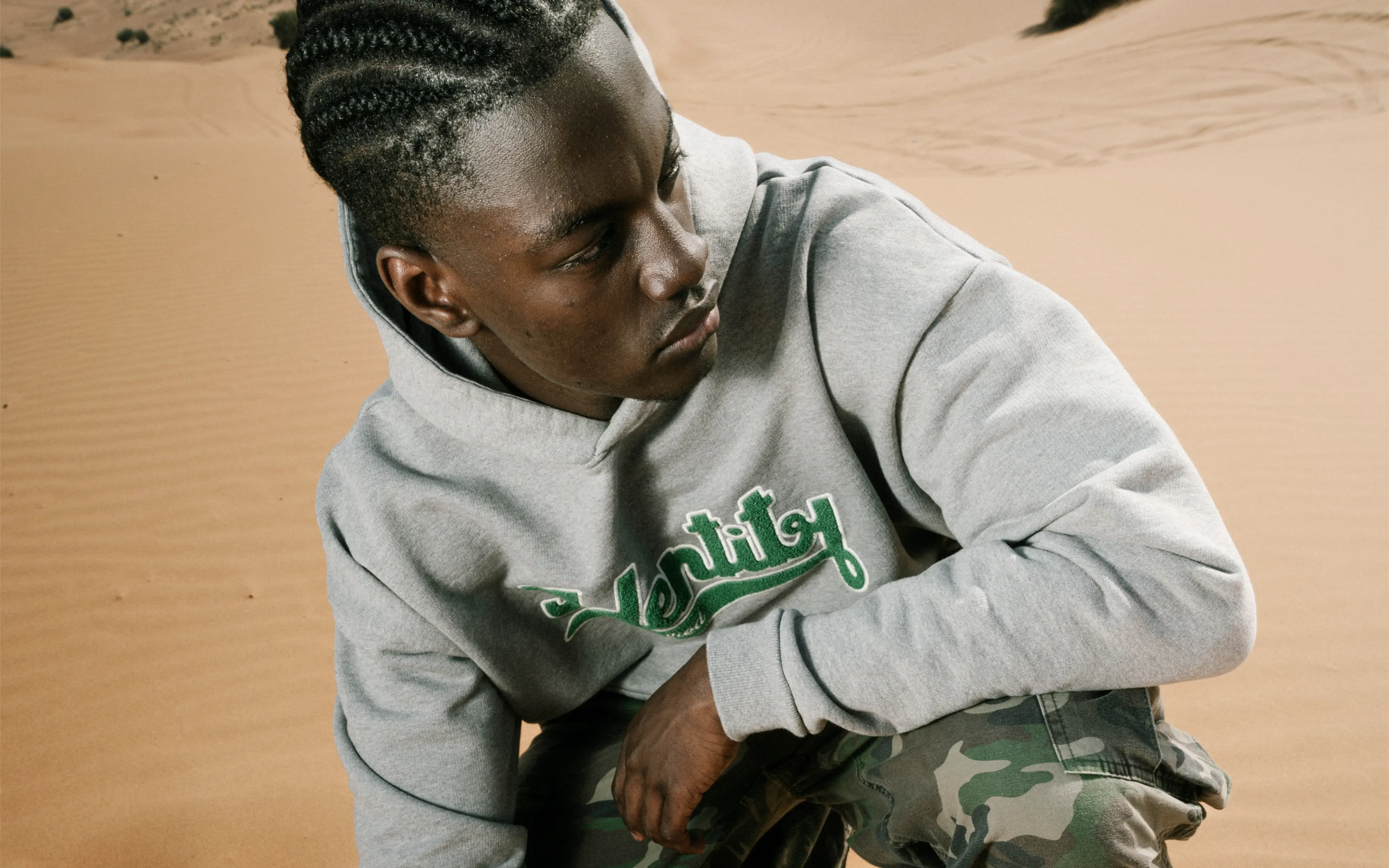 Ibo Ouro-Bodi sitzt im Sand und trägt einen grauen Certified-Identity-Hoodie mit grüner Stickerei.
