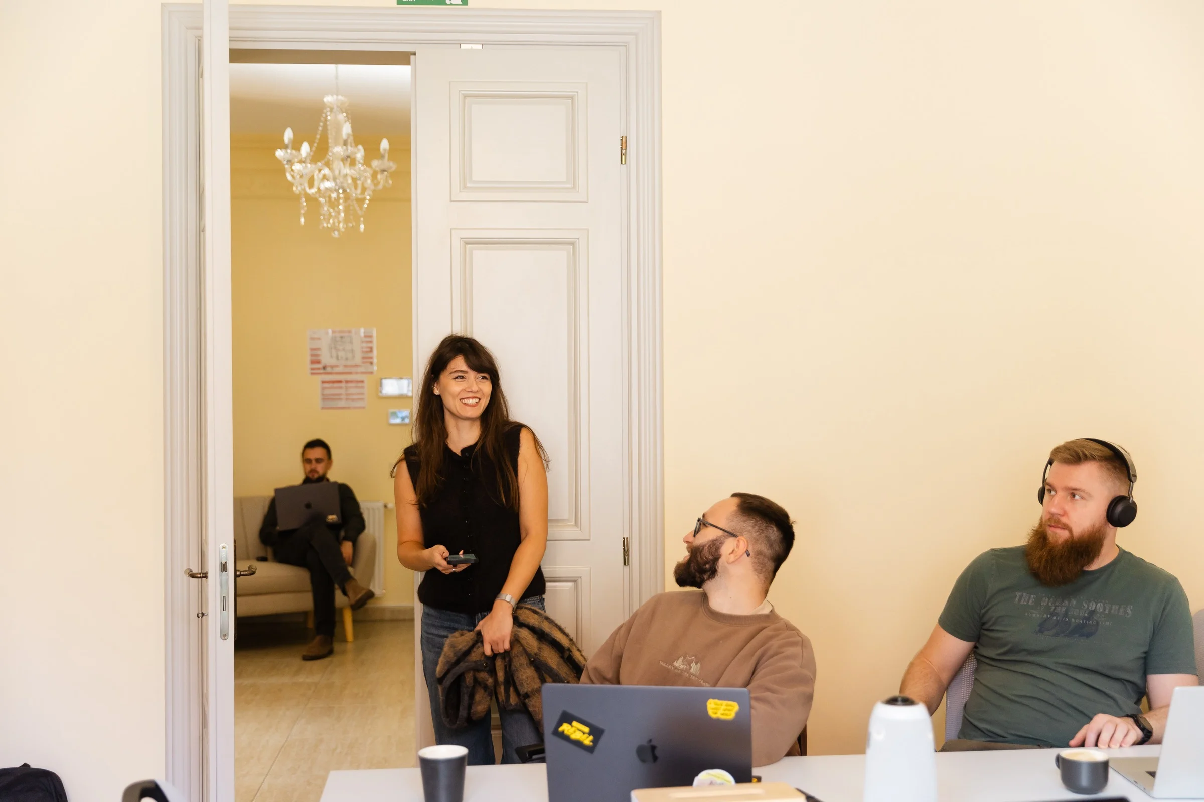 Three men and one woman in an office setting, with two men sitting at a table with laptops and the woman standing smiling near a doorway where another man sits on a chair with a laptop.
