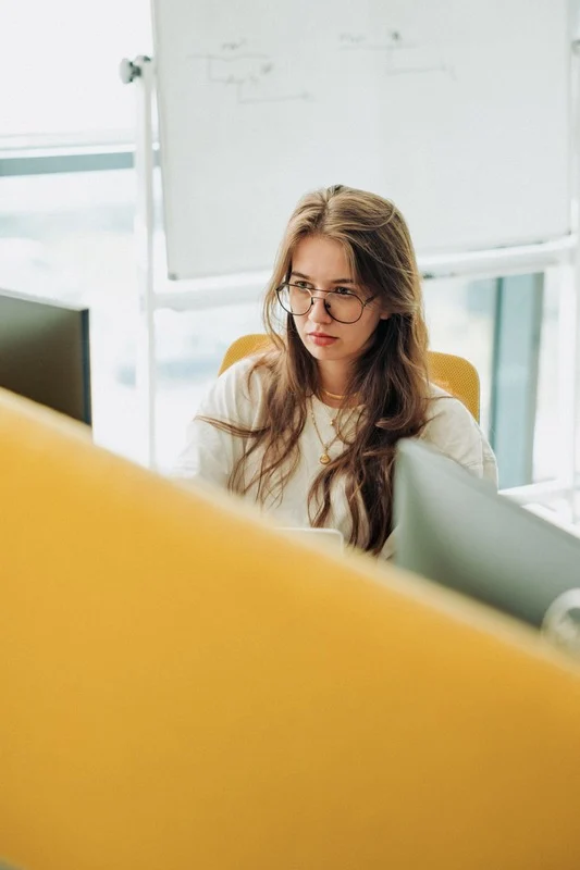Young woman with glasses working at a desk in a modern office with partitions and a whiteboard in the background.