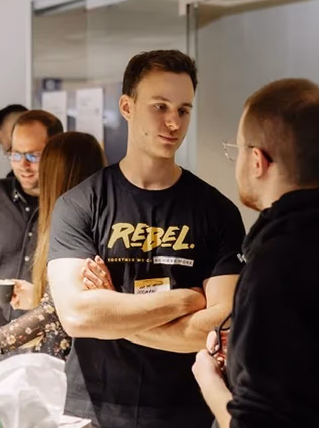 Young man with folded arms wearing a black T-shirt with the word 'REBEL' talking to another person in a casual indoor setting.