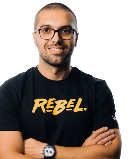 Mand med briller iført en sort t-shirt med ordet 'REBEL.' trykt på den, smilende med krydsede arme.