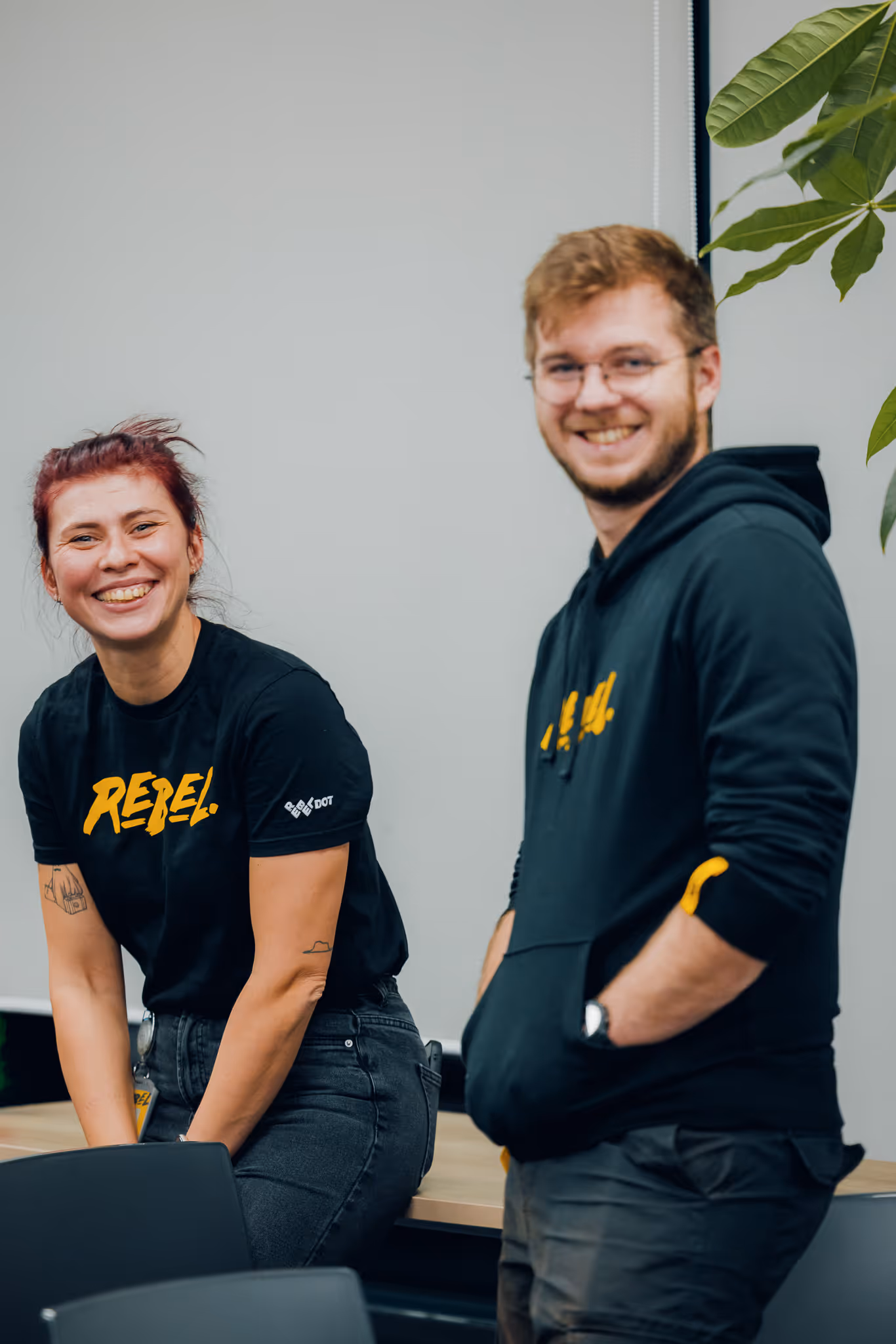 A smiling woman with red hair wearing a black REBEL t-shirt and a man with glasses wearing a black REBEL hoodie standing indoors near a table.