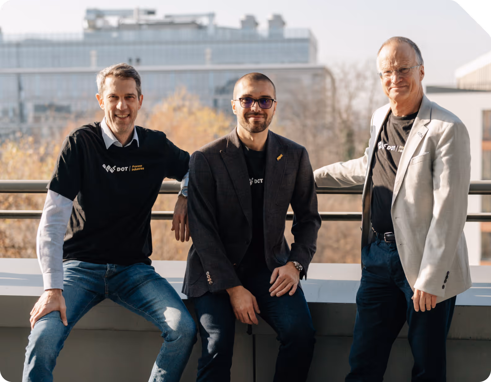 Three men smiling, with two wearing black DOT Process Industries t-shirts and one in a light blazer, sitting and standing outdoors with a cityscape background.
