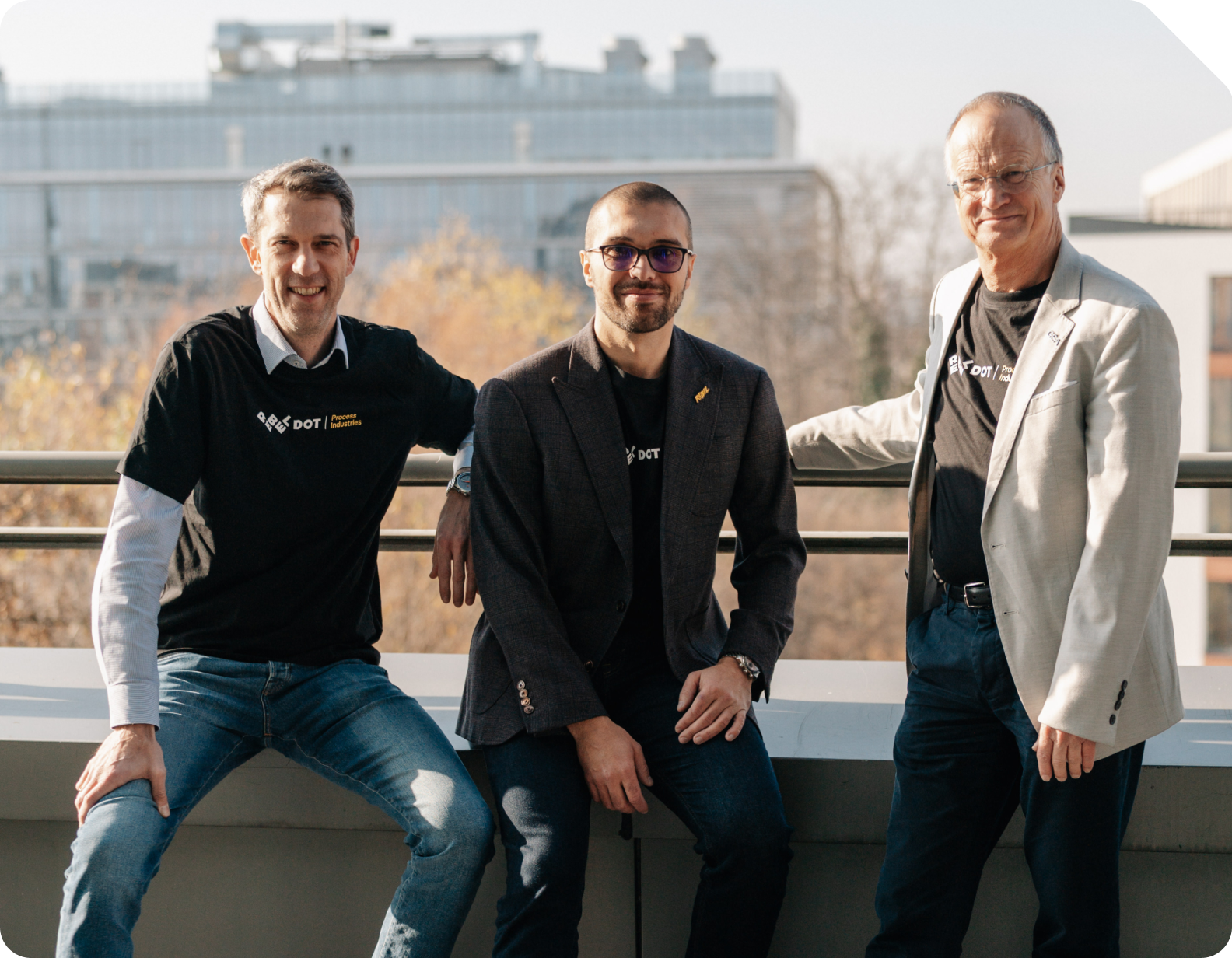 Tre mænd smilende, med to iført sorte DOT Process Industries-t-shirts og en i en lys blazer, siddende og stående udendørs med en bybilledbaggrund.