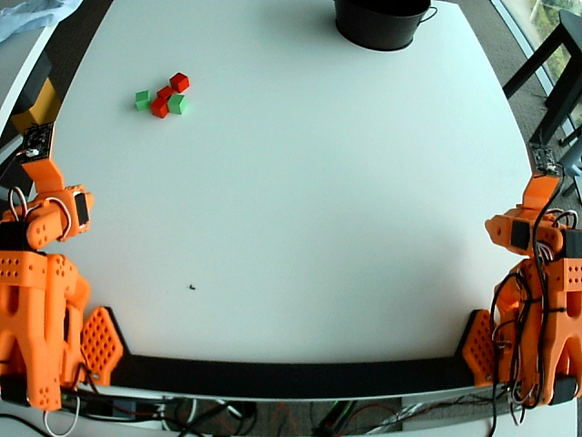 Top-down view of the table: two orange robot arms, red and green cubes in the upper left, black bucket in the top right.