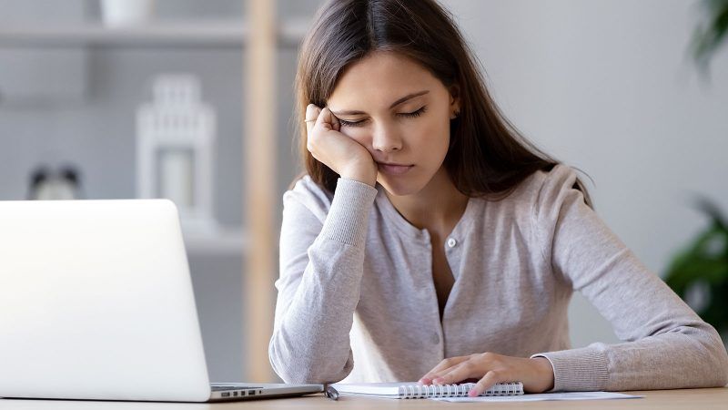 A woman sits at her laptop with her head in one hand and her eyes closed. She looks exhausted and bored.