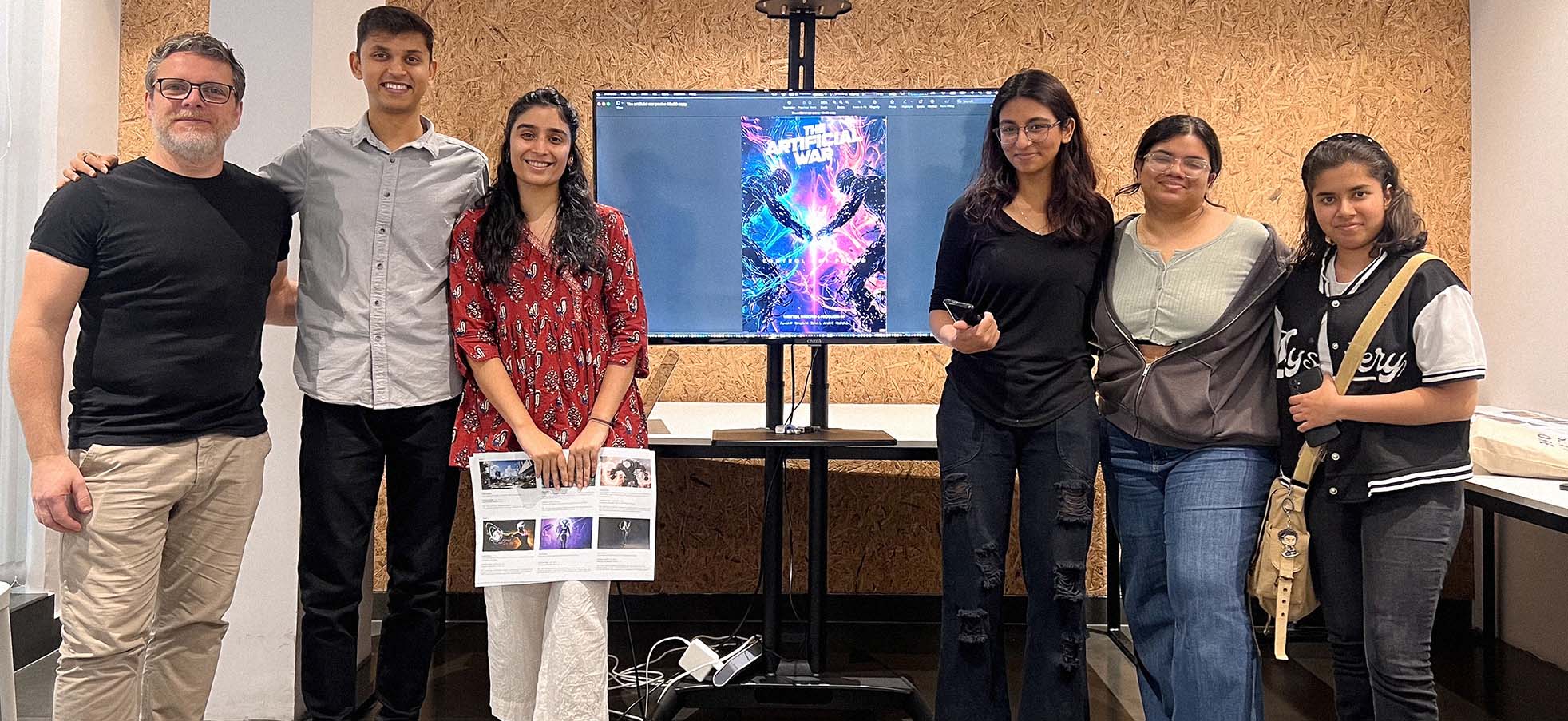Group of six design students standing in front of a screen displaying a vibrant poster titled 'The Artificial War' with two robotic figures facing each other.