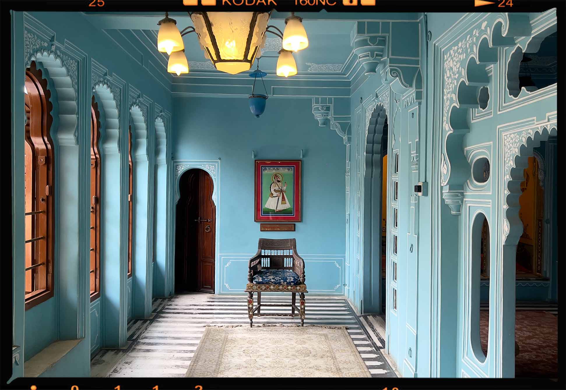 Intricately decorated blue-walled room with arched windows, a wooden chair with patterned cushion, and framed artwork on the wall.