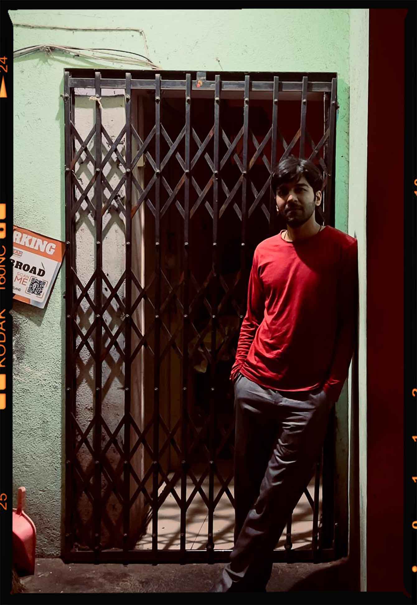 A man in a red long-sleeve shirt and gray pants leaning against a light green wall next to a closed metal gate at night.