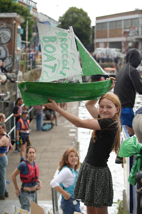 A young girl smiles while holding up a handmade green model sailboat with a sail that reads “BROCCOLI BOAT.” She stands by a busy dockside during what appears to be a community or festival event, surrounded by people in life vests and casual clothing.