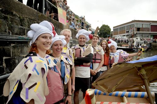 A group of people in whimsical costumes resembling ice cream sundaes smile beside a handmade boat. Dressed in sprinkle-covered capes and hats topped with cherries, they stand by the waterfront as crowds gather in the background for a festive event.