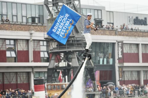 A performer in a sailor outfit holds a large blue "Bristol Harbour Festival" flag while soaring above the water on a flyboard. Jets of water propel him into the air as a crowd watches from the harbourside, with the M Shed museum visible in the background.