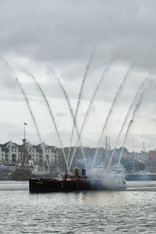 A vintage fireboat sprays powerful arcs of water into the air during a harbor display. Two crew members in high-visibility gear stand on deck as mist and smoke rise around them. In the background, historic ships and waterfront buildings line the edge of the harbor under an overcast sky.