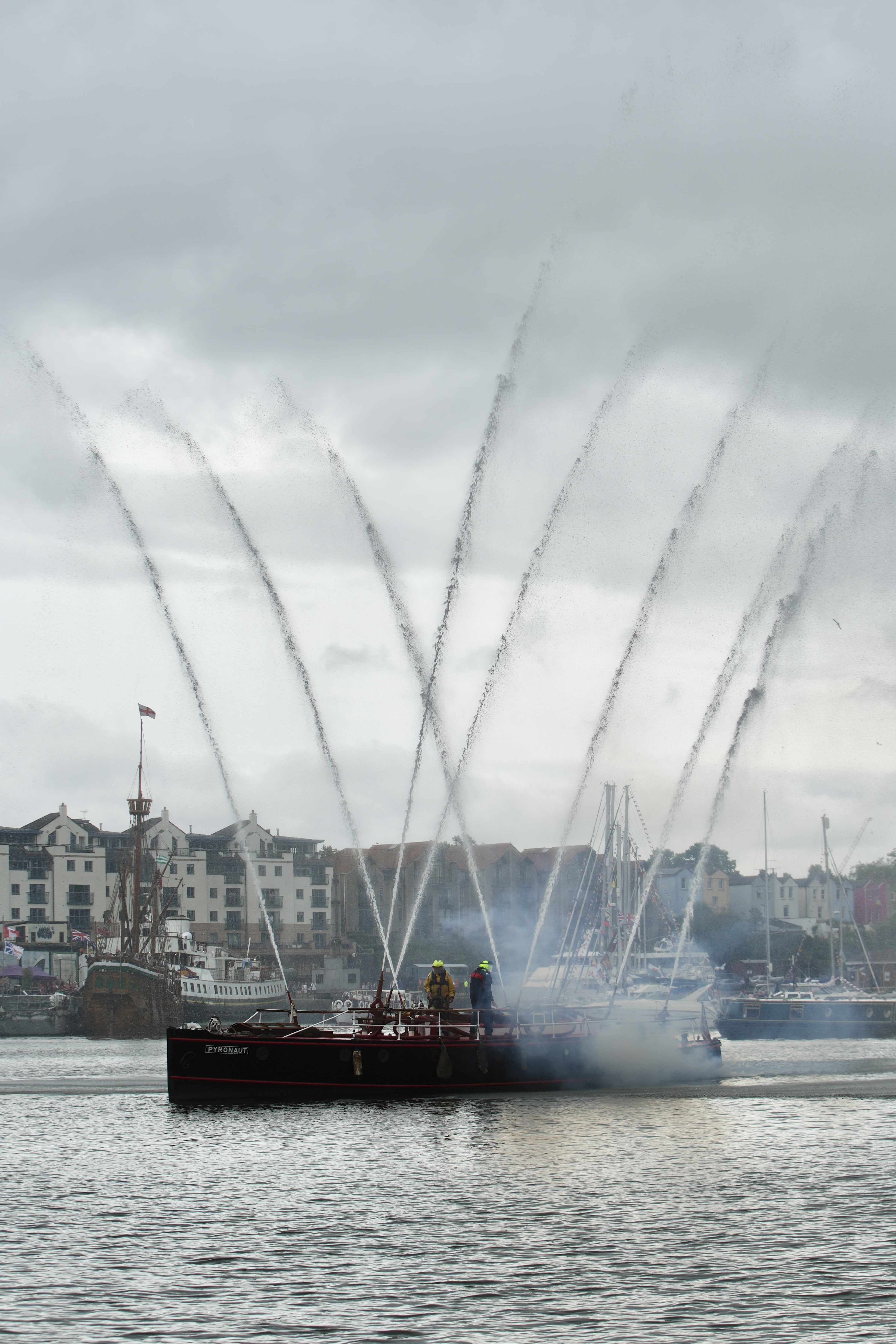 A fireboat sprays multiple high jets of water into the air in Bristol’s harbour, creating dramatic arcs against a grey, cloudy sky. Behind the boat, historic ships and colourful harbourside buildings are visible.