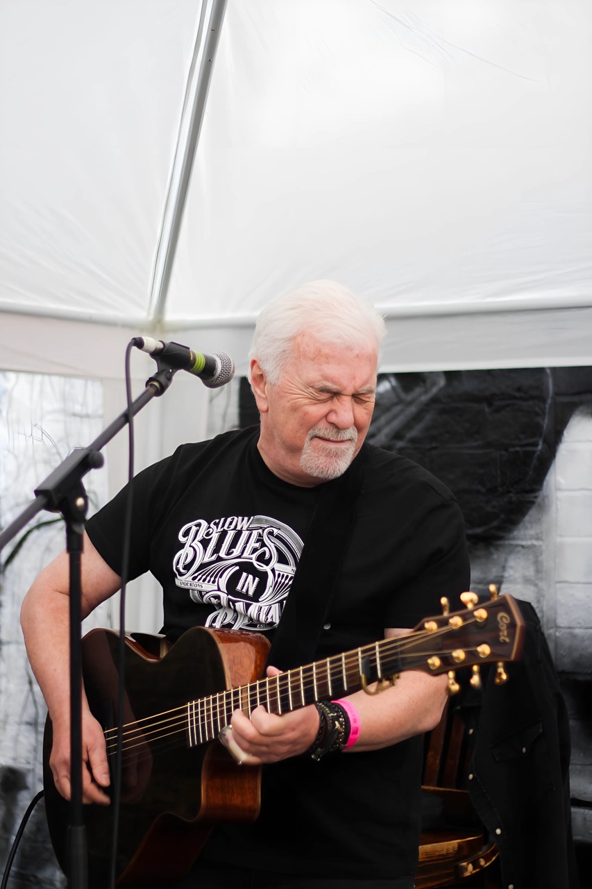 An older man with white hair and a goatee passionately plays an acoustic guitar under a white tent. Wearing a black “Slow Blues In” t-shirt, he performs with his eyes closed and a look of deep emotion, fully immersed in the music. A microphone and amplifier are set up in front of him.