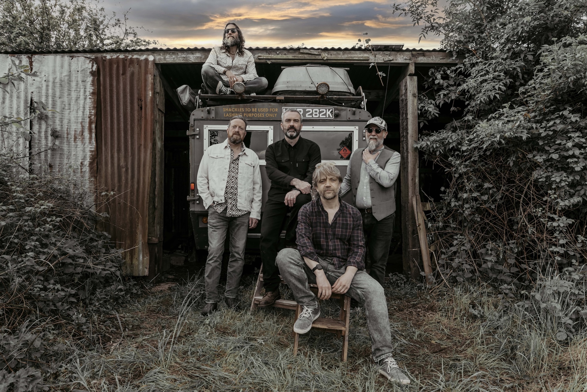 Six men sit on rustic wooden steps outdoors, framed by open wooden gates and surrounded by tall grass and overgrowth. The group, dressed in casual and rugged clothing, pose under a dramatic, cloud-filled sky, creating a moody and atmospheric portrait with a reflective tone.
