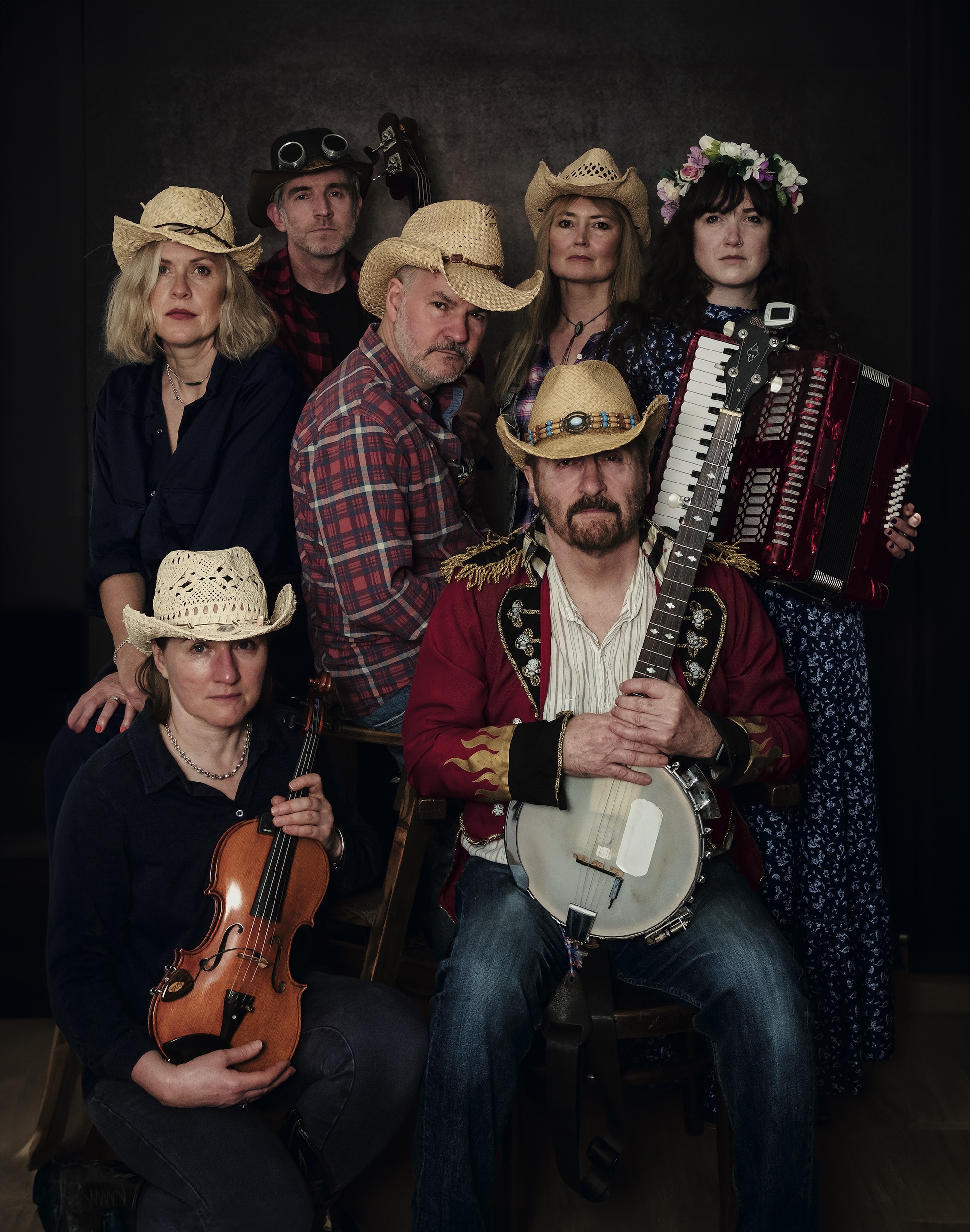 A group of seven musicians pose for a portrait in Americana-style outfits, wearing cowboy hats, flannel shirts, and floral accessories. They hold instruments including a banjo, violin, accordion, and upright bass, giving off a rustic, old-time country band vibe with serious, focused expressions.