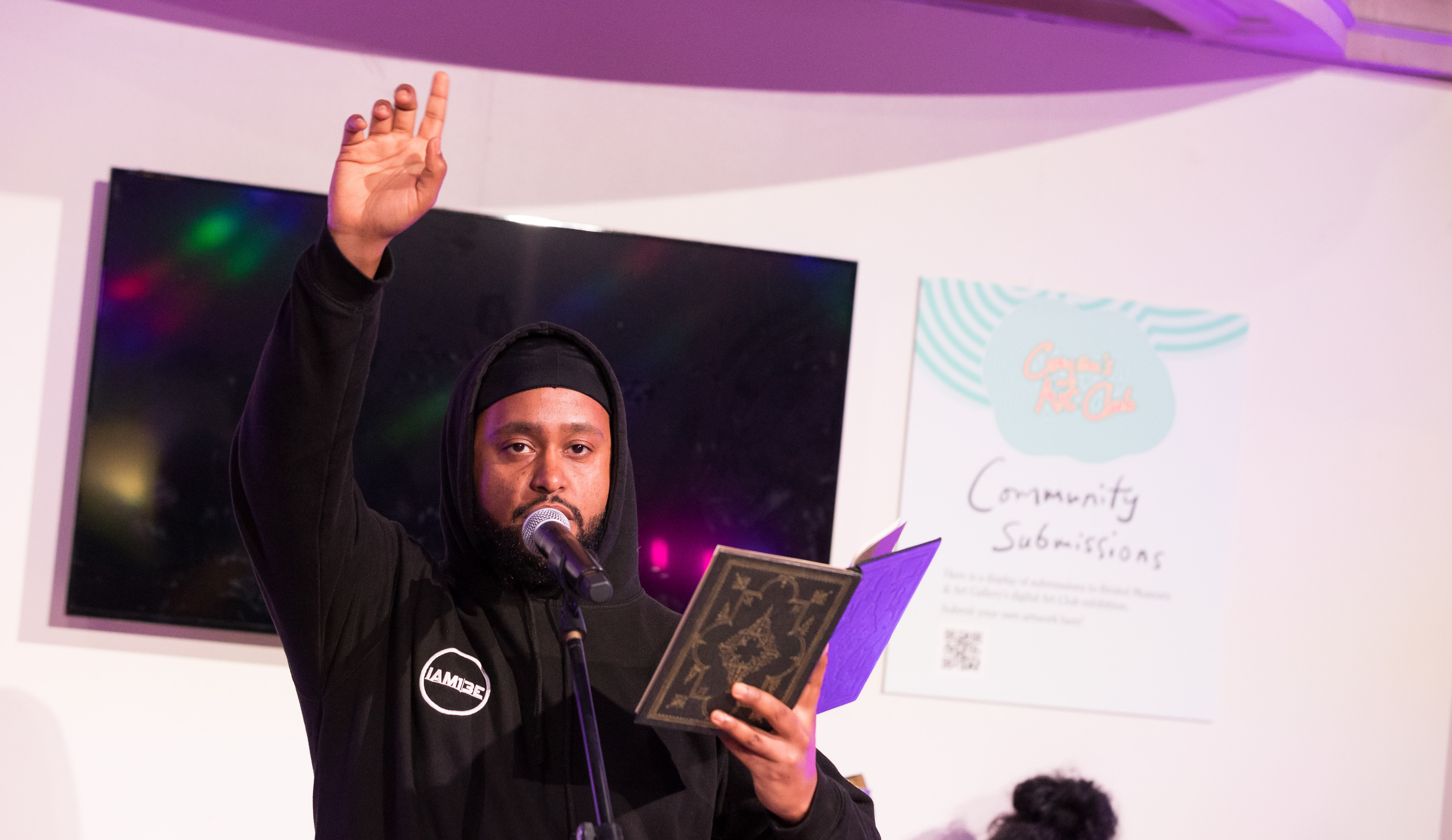 A man in a black hoodie stands at a microphone, holding an open book and raising one hand. He appears to be performing or reading aloud in a brightly lit indoor space.