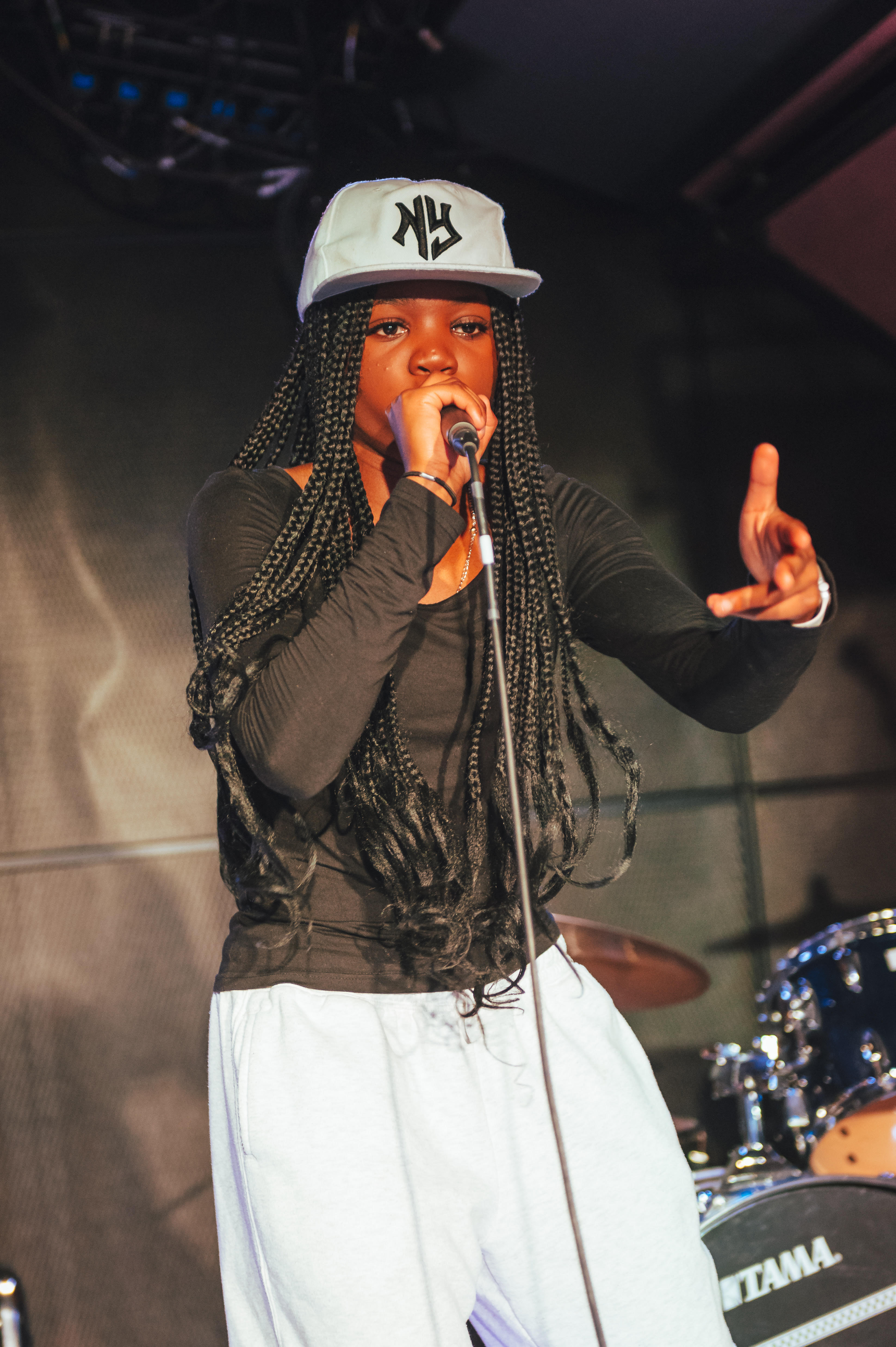 This powerful image captures a young performer mid-flow, confidently holding the mic with one hand while gesturing expressively with the other. Dressed in a black top, white sweatpants, and a New York-style cap, she delivers her lyrics with intensity and focus. The stage lights highlight her presence, with a drum kit softly lit in the background, hinting at a live music setting full of energy and promise.