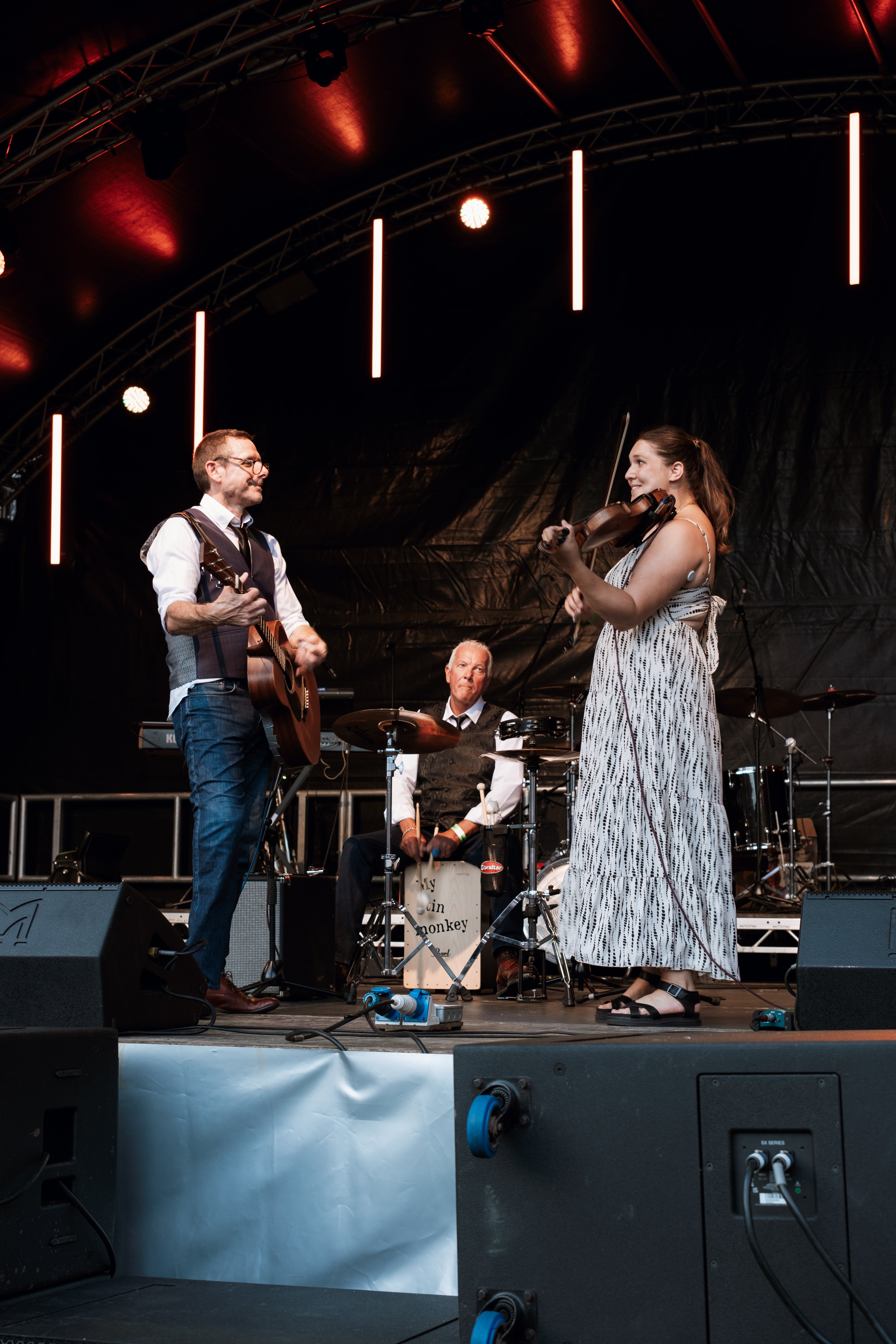 A three-piece band performs on stage under warm lighting. A man plays acoustic guitar and sings, a woman in a patterned dress plays the violin, and a third member sits at a drum box labeled “My Tin Monkey.” The group appears energetic and engaged, with a dark backdrop and professional sound setup behind them.