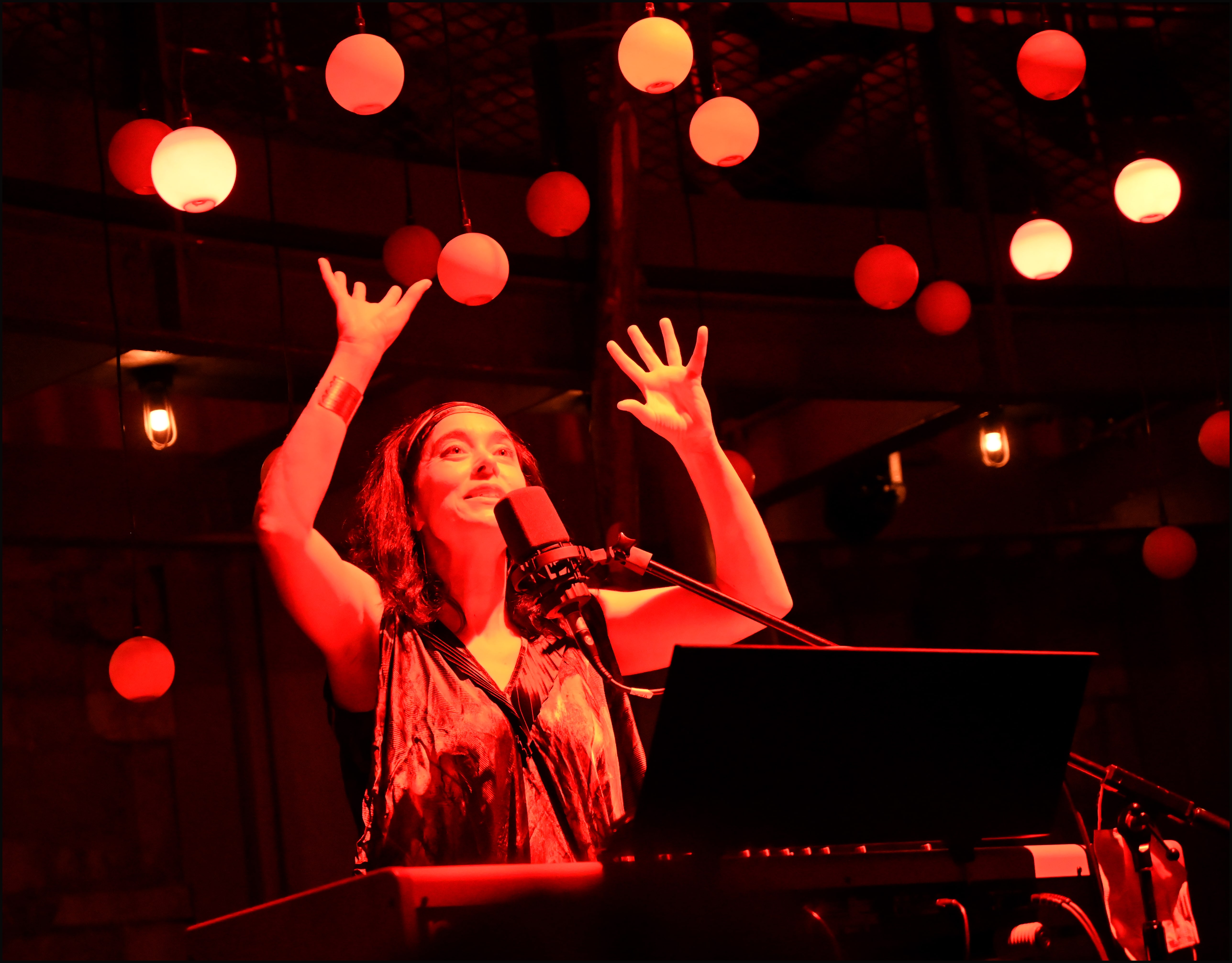 A vibrant, red-lit image captures a woman mid-performance, standing behind a keyboard and microphone. With her hands raised expressively, she looks upward, surrounded by an array of glowing, suspended orbs. The setting suggests an immersive and atmospheric live show.