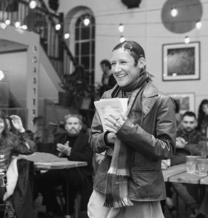 This candid black-and-white image captures a joyful moment during what appears to be a spoken word or literary event. The subject, smiling warmly and holding a stack of papers, stands confidently among a crowd of engaged and supportive audience members. The cozy, creative atmosphere is enhanced by string lights, framed artwork, and the visible word "ROASTER" hinting at a café setting. The energy feels inclusive, expressive, and full of heart.