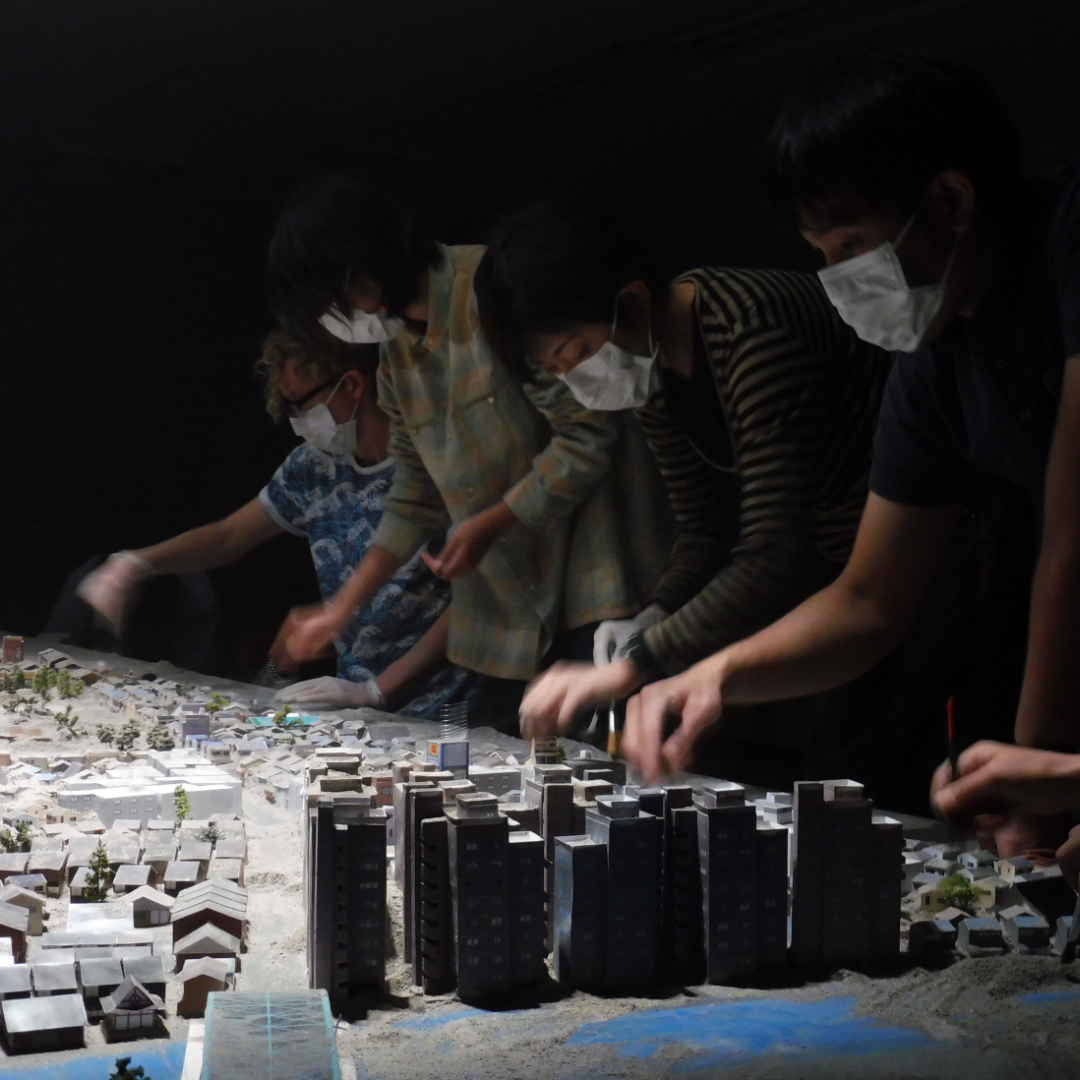 This image shows a group of people, all wearing face masks, carefully brushing or working on a large-scale, highly detailed model of a cityscape. The dim lighting and the participants' intense focus give the scene a solemn, almost ceremonial tone. It suggests themes of preservation, remembrance, or restoration—perhaps connected to a historical event or urban memory project. The presence of dust or ash on the model adds a poignant, reflective layer to the activity.