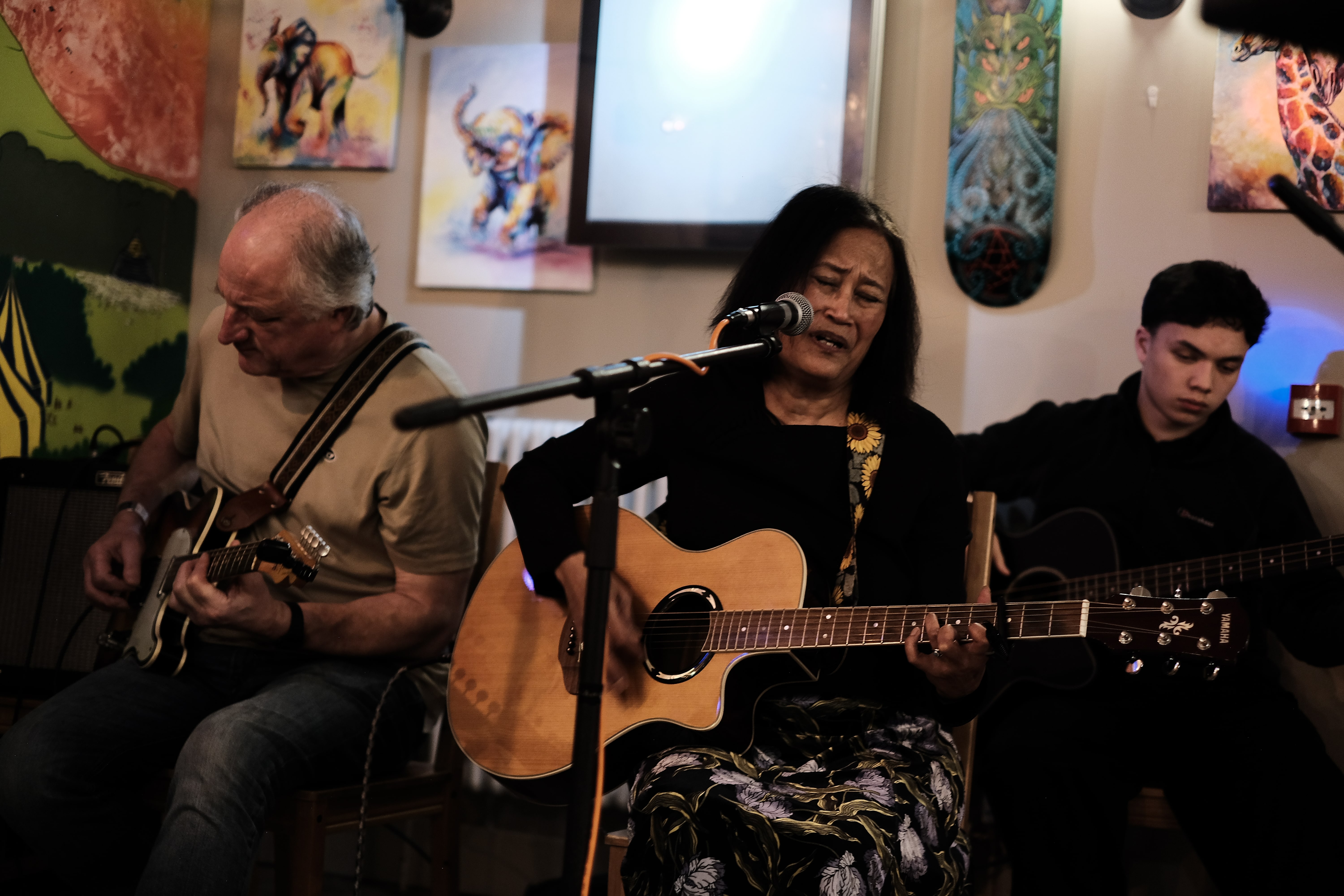 This image captures an intimate live music performance, featuring three musicians deeply engaged in playing acoustic and electric guitars. The central figure—a woman singing with closed eyes conveys strong emotion, suggesting a heartfelt or soulful performance. The setting appears cozy and creative, with vibrant paintings and eclectic decor like a painted skateboard and animal-themed artwork on the walls, hinting at a community or arts café vibe. The overall scene reflects a warm, supportive atmosphere for local or grassroots musical expression.