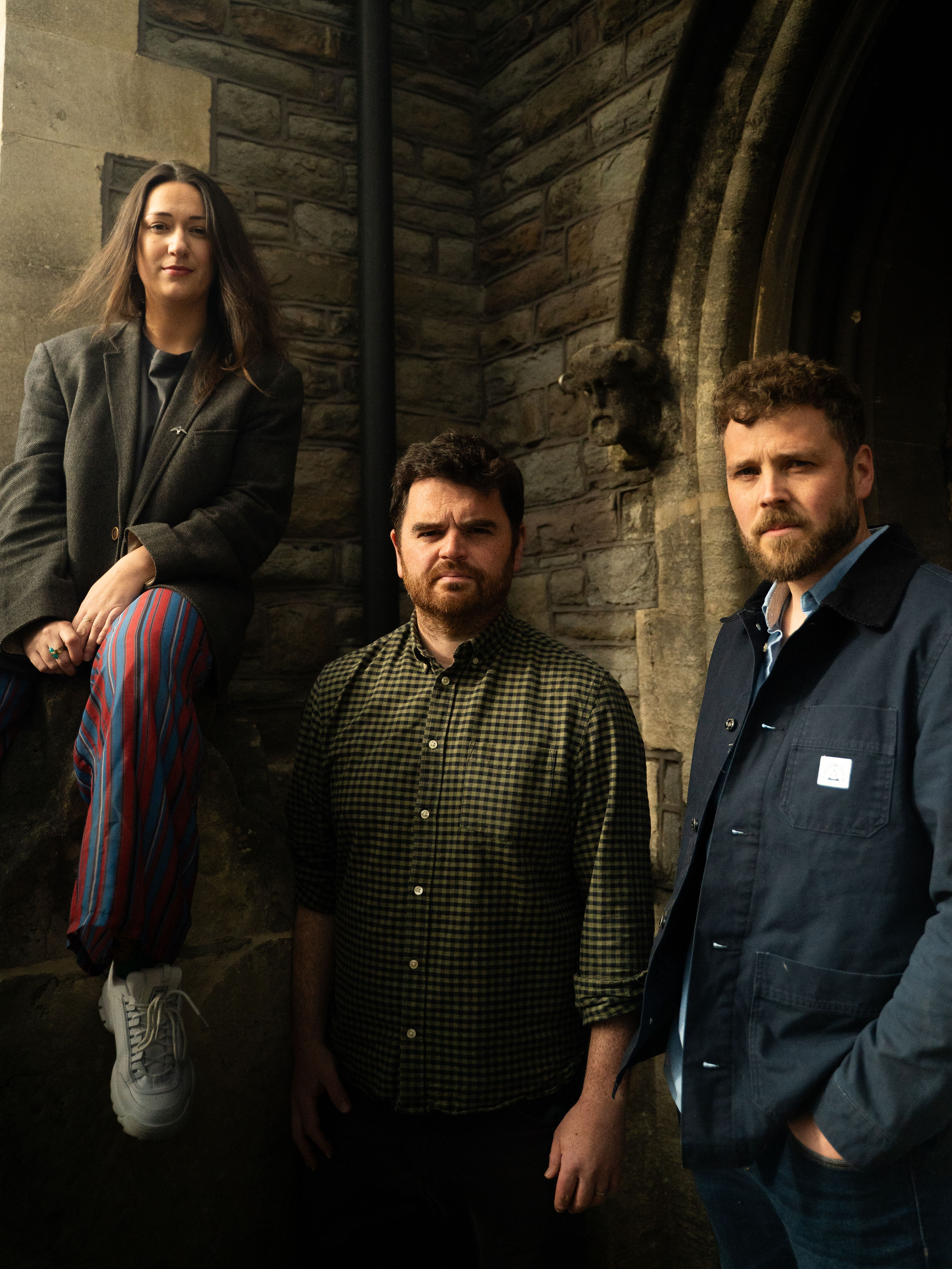 This striking image features a trio standing against a historic stone backdrop, likely the entrance of a church or heritage building. The lighting casts a warm, cinematic tone across their expressions and attire.