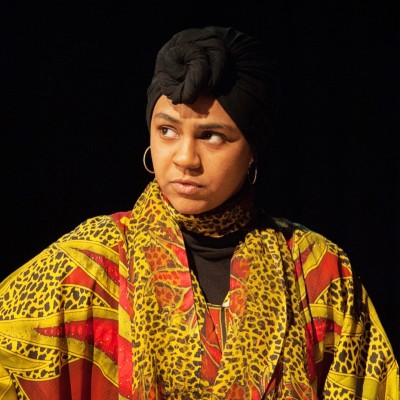 This image features a powerful portrait of a woman in bold patterned attire, including a striking red, yellow, and black shawl and a black headwrap. Her expression is focused and commanding, set against a dark backdrop.