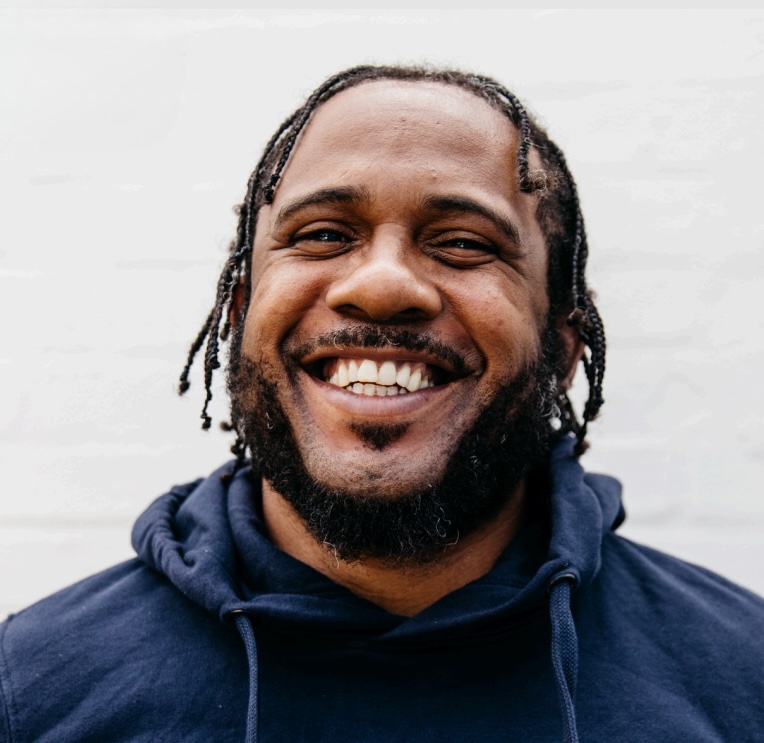 This image features a smiling man with braided hair and a beard, wearing a navy hoodie. His warm, confident expression is set against a clean, white background.
