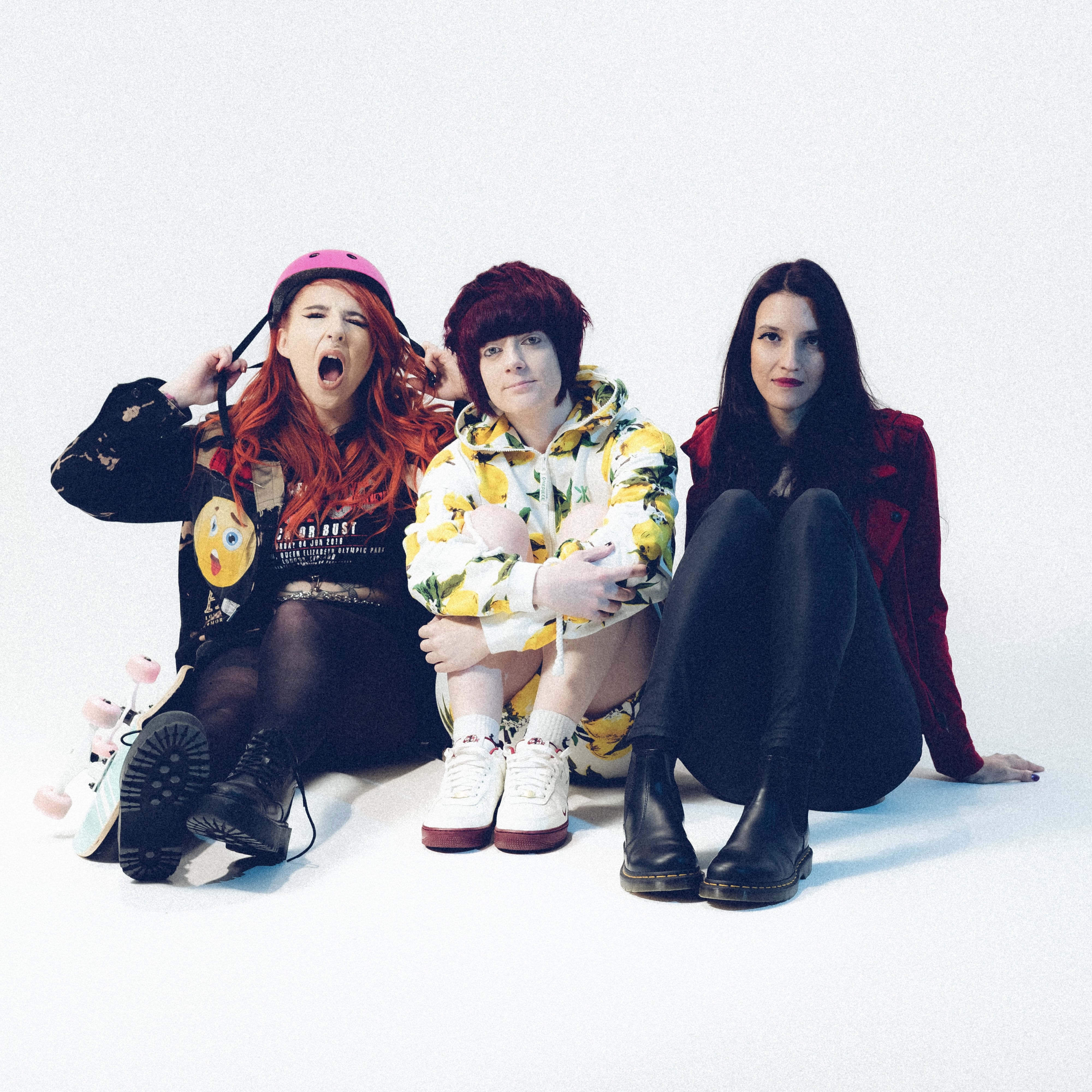 Three women sit on the floor against a white backdrop. The woman on the left wears a pink helmet, has bright red hair, and holds a skateboard. The woman in the middle wears a matching lemon-print hoodie and shorts set, and the woman on the right wears black leggings and a red jacket. All three pose casually, exuding a playful and bold energy. Ask ChatGPT