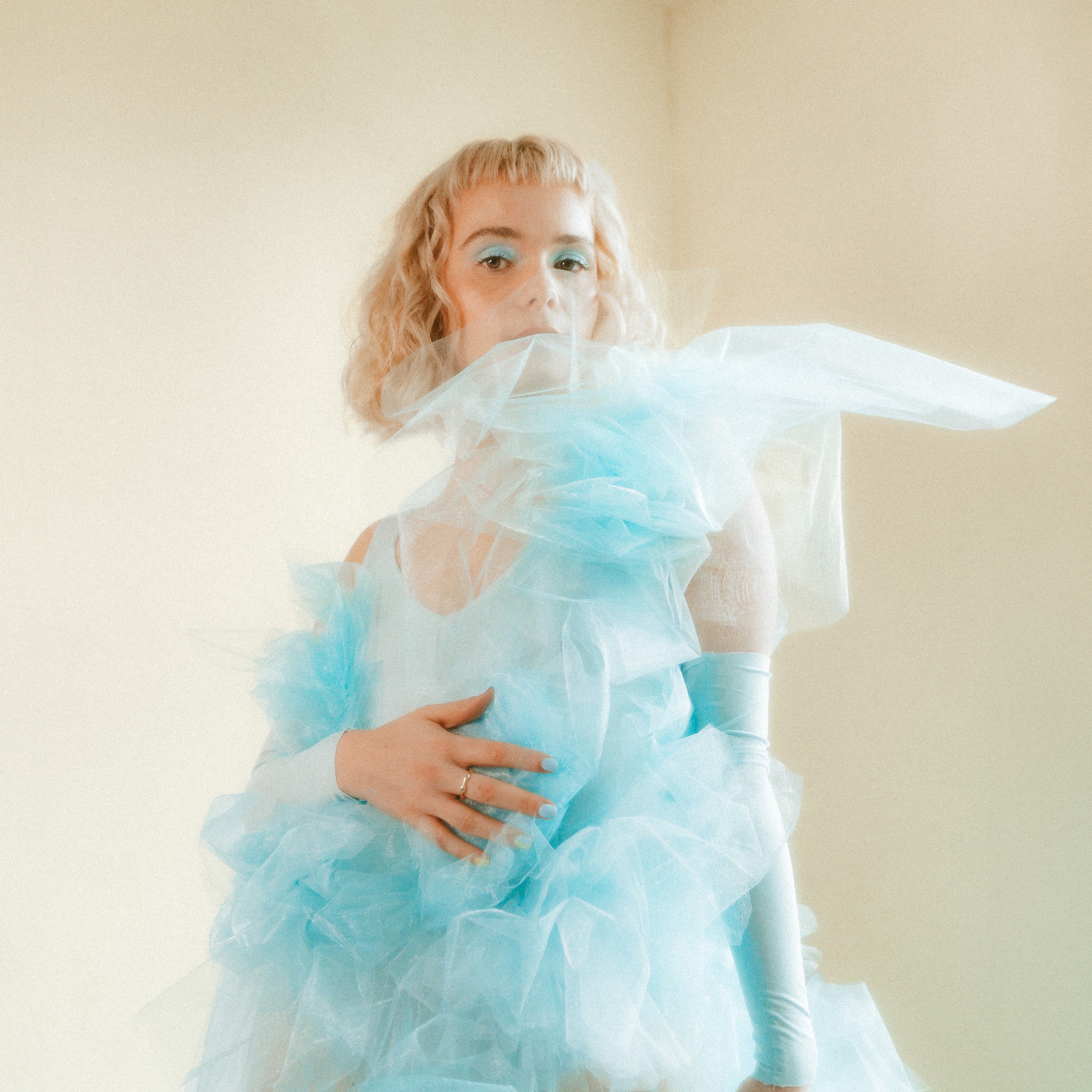 A woman with short blonde curls and pastel blue eyeshadow poses in a soft-lit room wearing a dramatic, cloud-like blue tulle outfit. Her hand rests gently on her chest, matching her pastel blue nail polish to the flowing fabric.