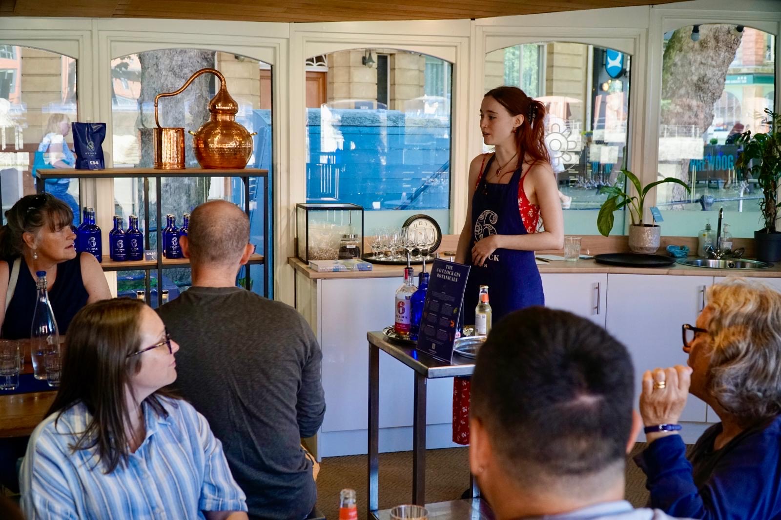 A young woman in an apron stands at the front of a small tasting room, speaking to a seated group of adults. In front of her is a table displaying gin bottles, glasses, and a menu. Behind her, shelves display blue gin bottles and a copper distilling pot. The setting is casual and inviting, with natural light streaming through large windows.