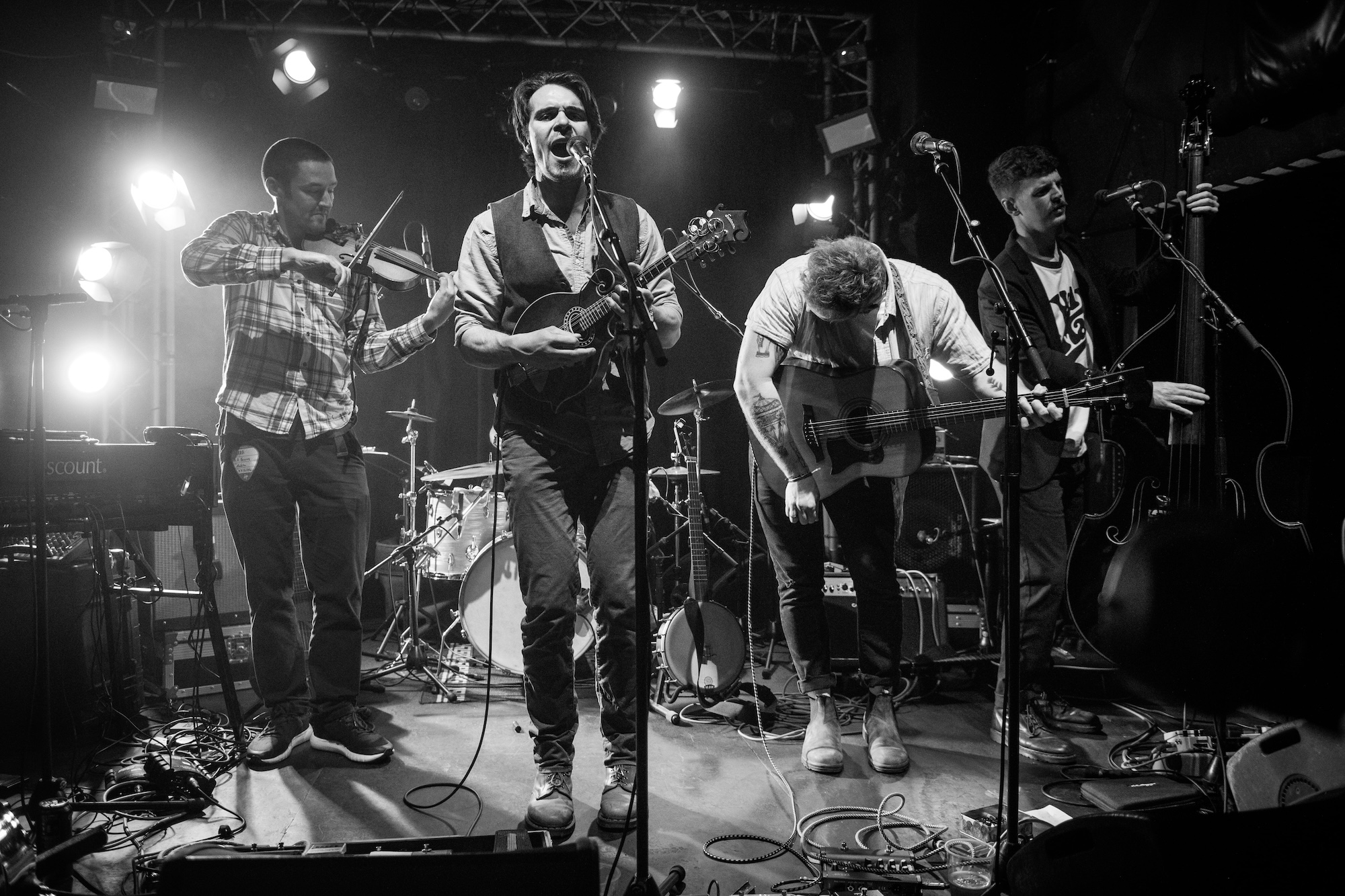 Black and white photo of a five-piece band performing on stage. The lead singer stands center stage playing a mandolin and singing passionately into the microphone. To his left, a violinist plays intently, and to his right, two guitarists—one mid-bow and the other adjusting his instrument. A double bassist stands at the edge. The stage is lit with concert lights and surrounded by musical equipment.