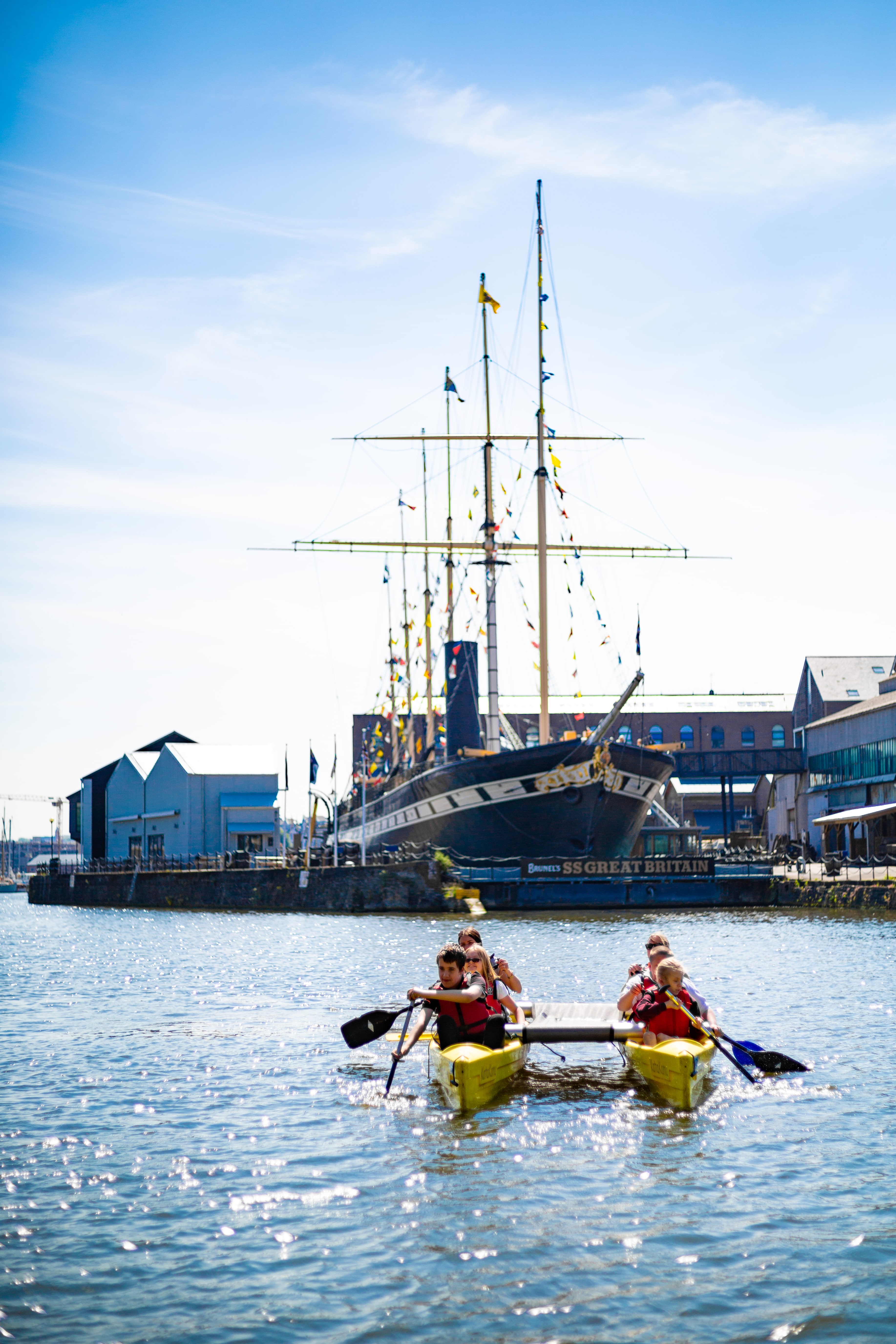 A group of people in life jackets paddle a yellow canoe-like catamaran across Bristol Harbour on a sunny day. Behind them, the historic SS Great Britain, adorned with colourful flags, is docked at the harbourside, with surrounding buildings and a clear blue sky completing the scene.