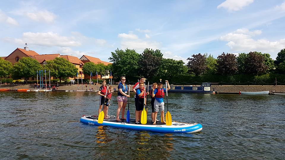 A group of five people wearing life jackets stand on a large paddleboard on a calm river, each holding a paddle and smiling. In the background, trees and a riverside building with a red roof are visible under a sunny, partly cloudy sky.
