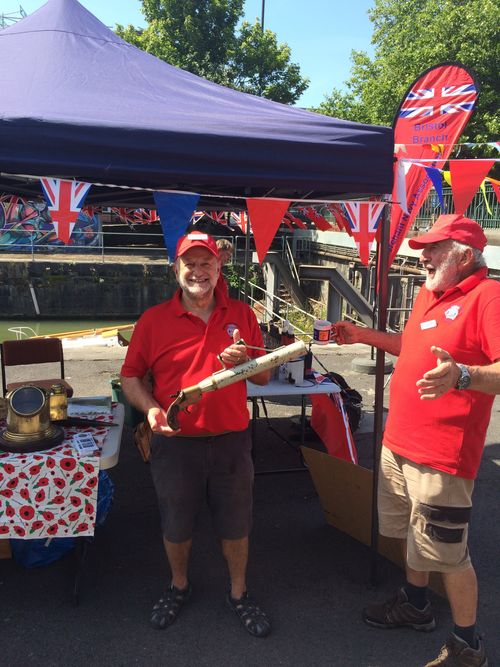 Two older men in red shirts and hats smile and chat at a heritage-themed outdoor stall decorated with Union Jack flags and bunting. One man holds an antique maritime object, while the table displays brass nautical instruments and poppy-themed decorations.