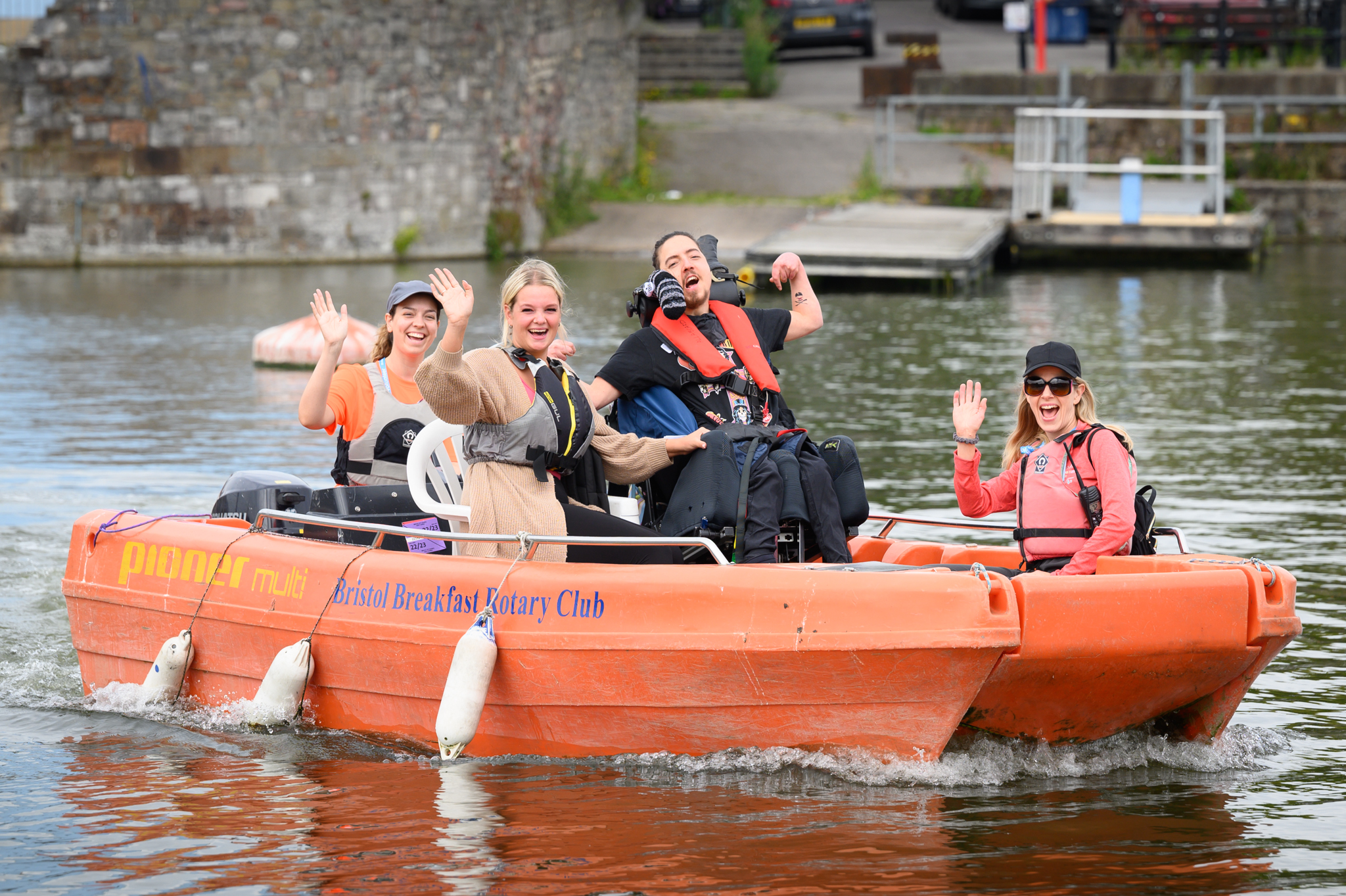 A group of five smiling people wave cheerfully from an orange motorboat labeled "Bristol Breakfast Rotary Club" as they ride across a calm body of water. One of the passengers is a wheelchair user, and everyone is wearing life jackets. A stone wall and dock are visible in the background.
