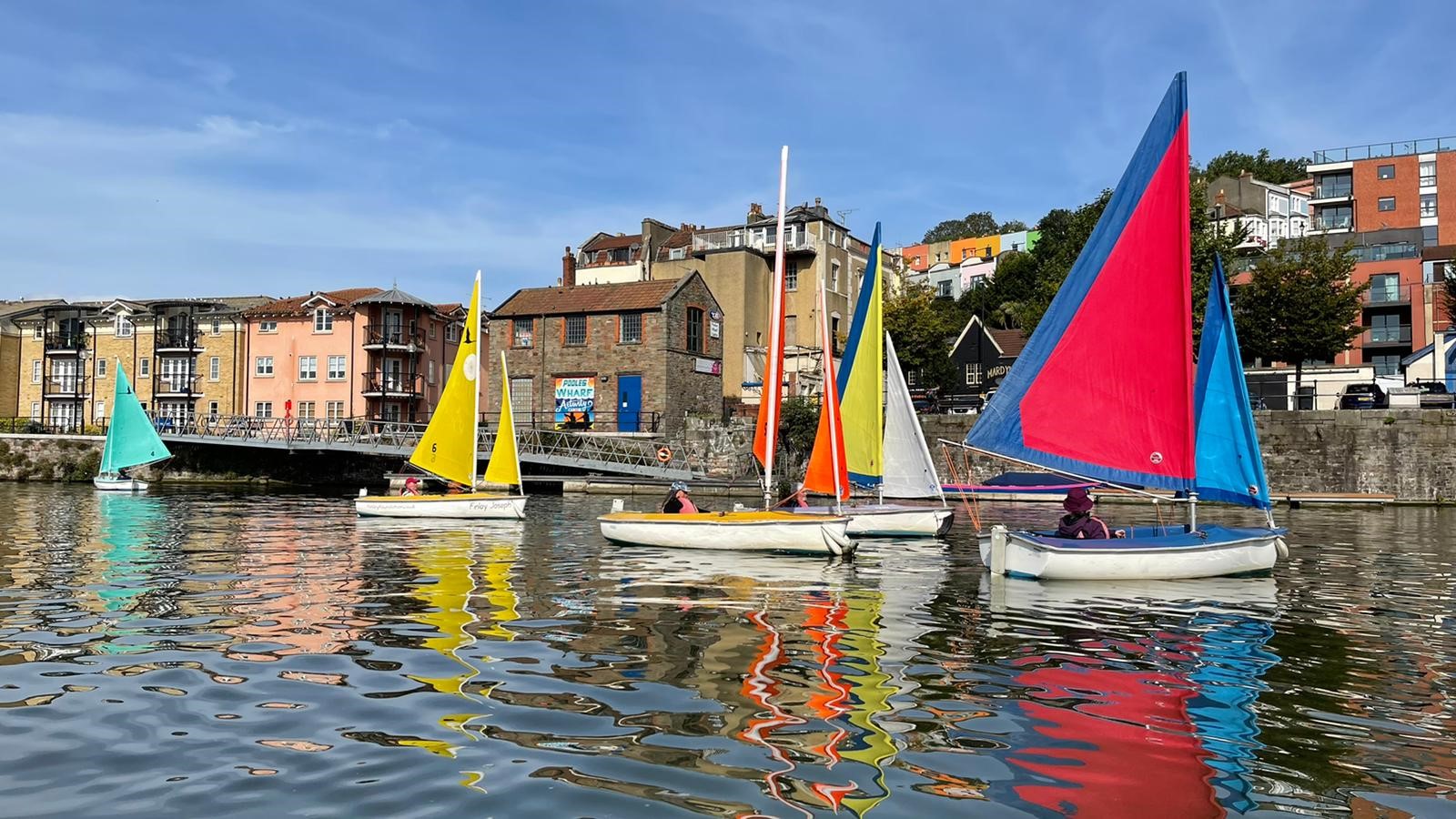 A colourful fleet of small sailboats with vibrant sails in shades of yellow, red, blue, and orange glides across the calm water of Bristol Harbour. The boats reflect vividly on the rippling surface. In the background, pastel-coloured buildings and harbourside architecture line the quay under a clear blue sky.