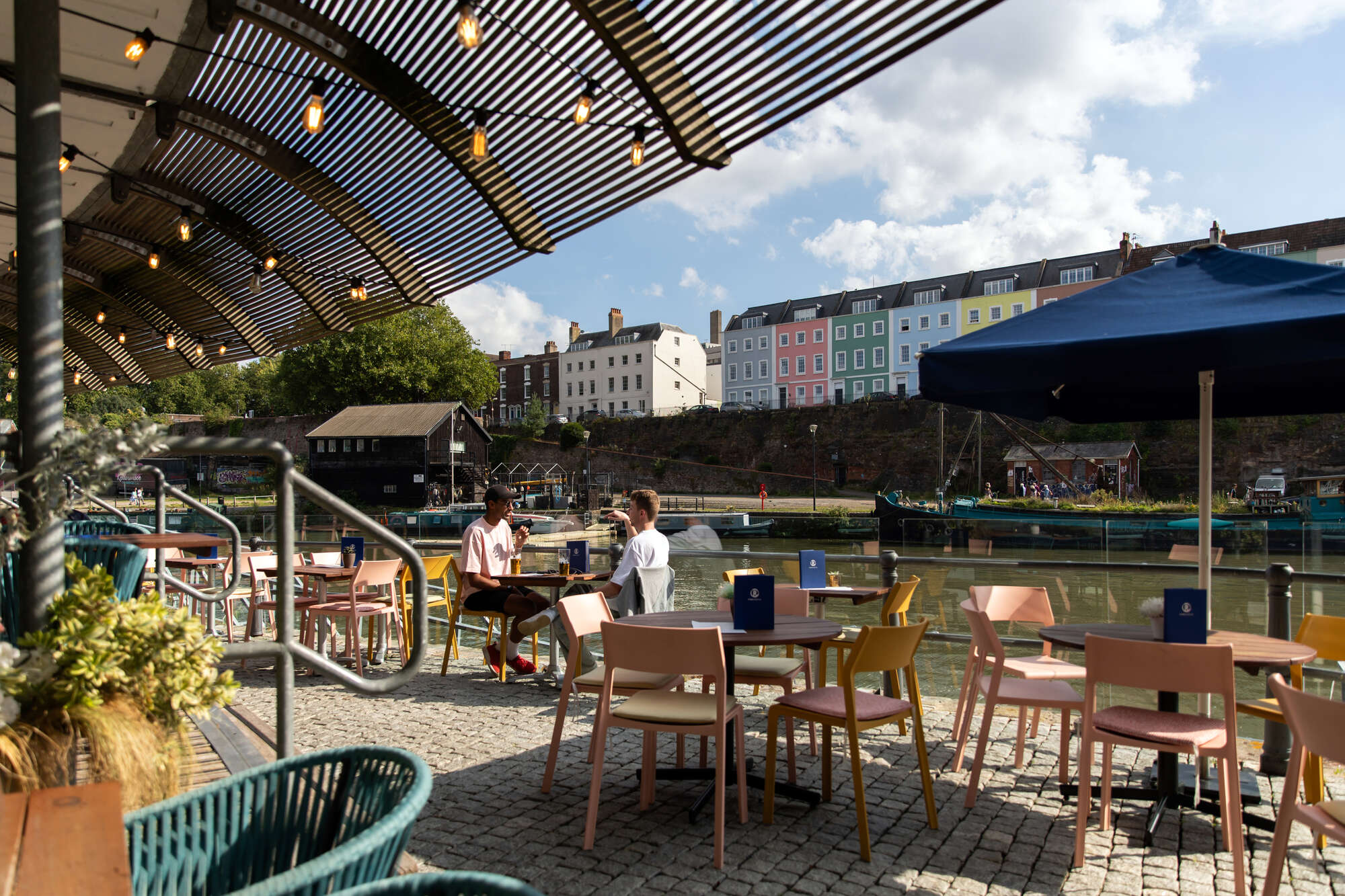 Two people sit at a waterfront café table on a sunny day, with pastel-colored houses on a hill and boats moored along the river in the background. The terrace is covered by a slatted canopy with hanging lights, and surrounded by modern pastel and wicker chairs.
