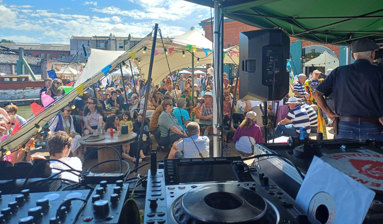 View from behind a DJ booth at a lively outdoor festival. A large crowd of people, including families with children, are gathered under white canopy tents decorated with colorful bunting. The event takes place by the water on a sunny day, with people seated at round tables enjoying the music and atmosphere.