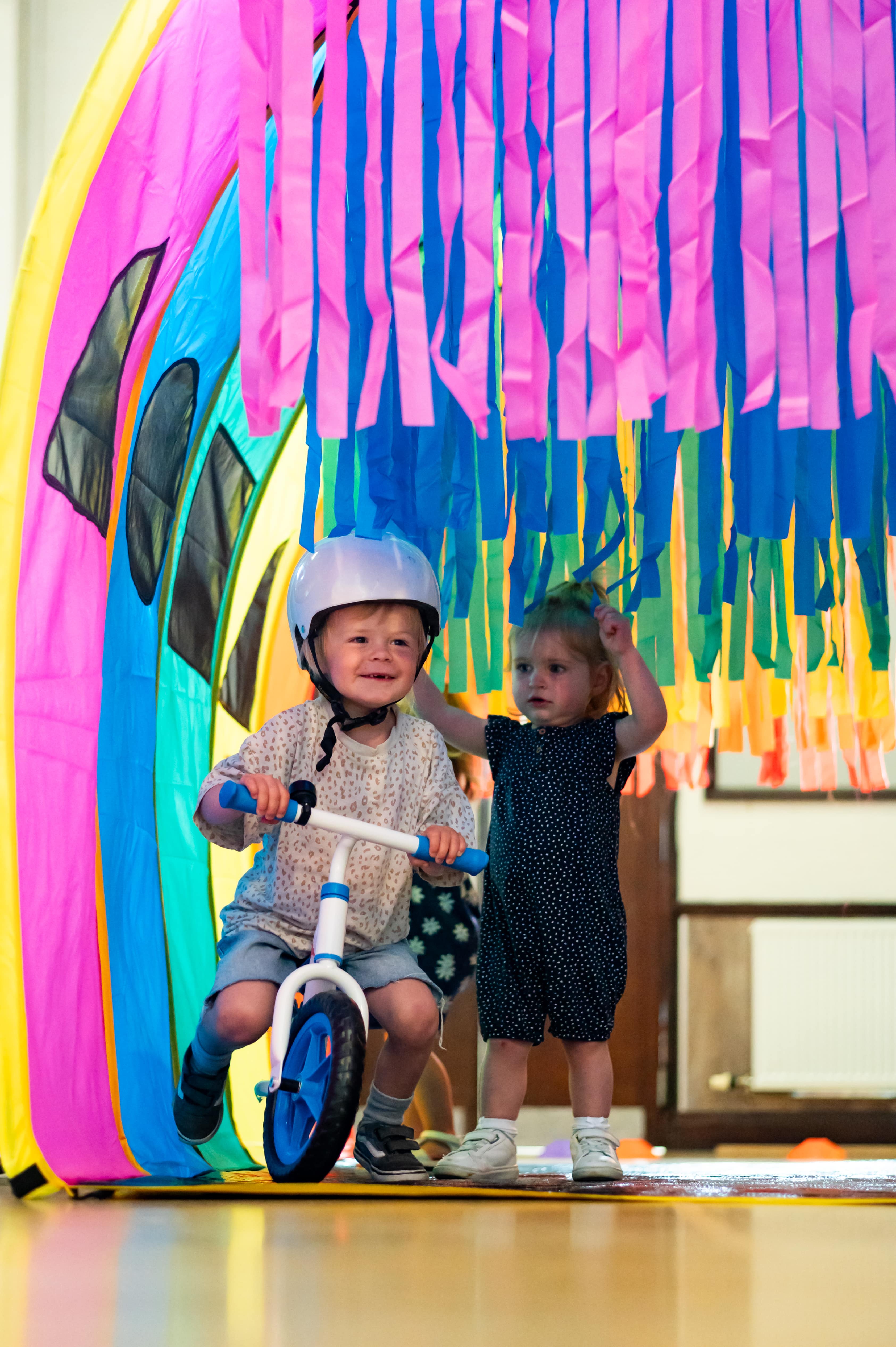 Two young children play under a rainbow tunnel decorated with hanging pink, blue, and green streamers. One child rides a balance bike wearing a helmet, while the other stands beside, reaching up to touch the streamers.