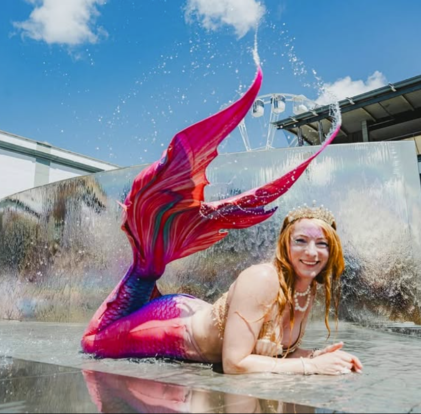 A smiling woman dressed as a mermaid poses in shallow water, wearing a vibrant pink and purple mermaid tail and a pearl crown. Water splashes around her as she lies in front of a shiny metallic backdrop under a bright blue sky.