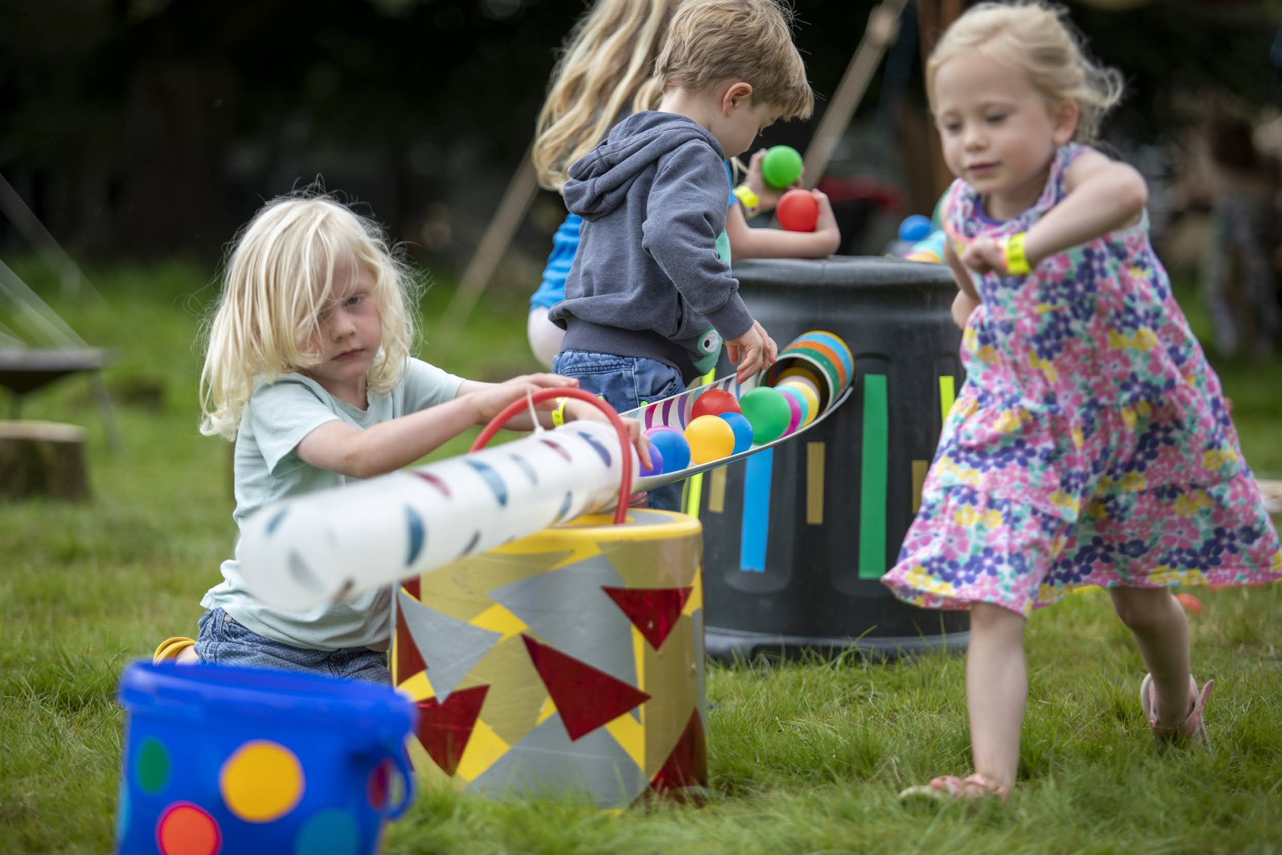 Children play with colourful balls and recycled tubes in an outdoor activity setup on grass. One child in a blue t-shirt places balls into a white tube decorated with red and blue dots, while other children move and explore around the interactive installation made of decorated barrels and materials.