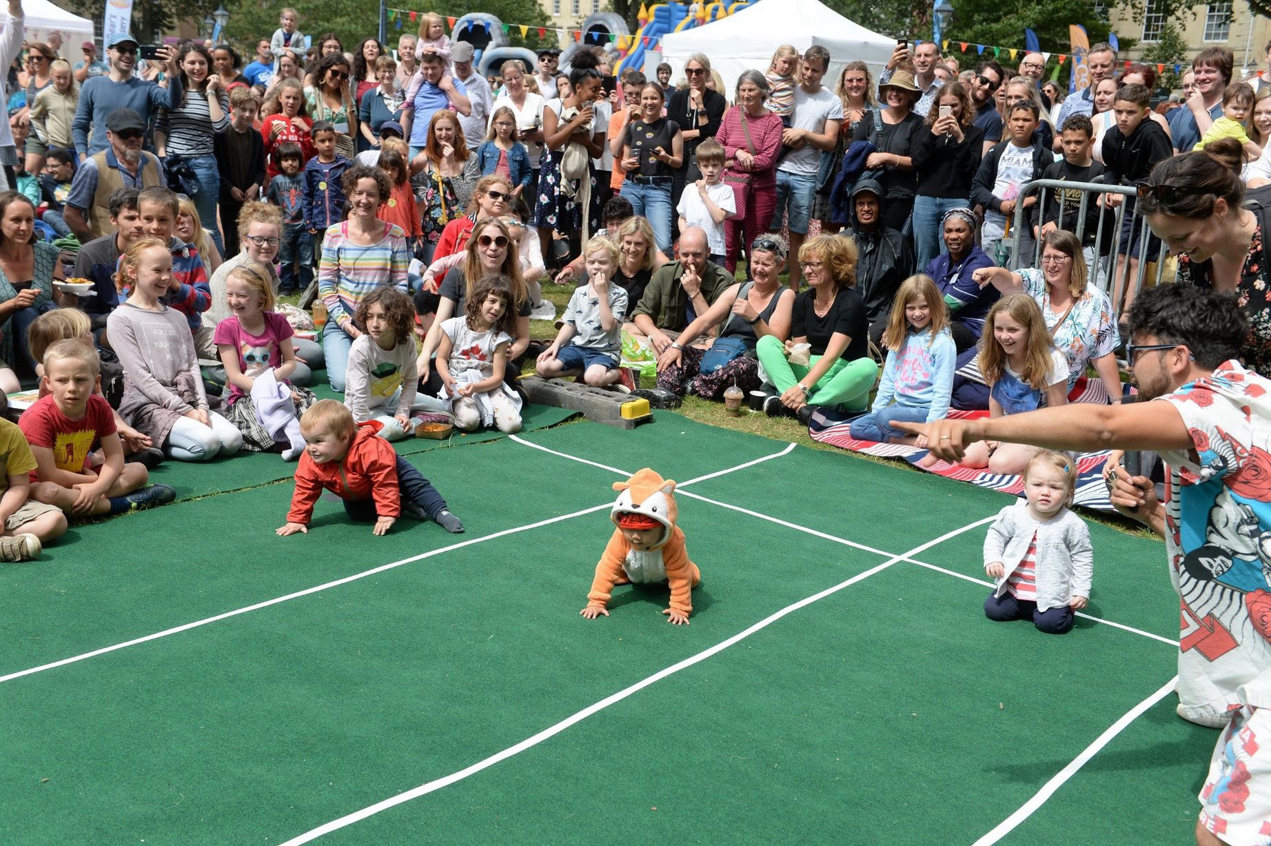 A large crowd of adults and children cheer and watch as three babies participate in a playful baby race on a green track with white lines. One baby is dressed in a fox onesie and crawling ahead, while others sit or crawl behind. A man in a colourful shirt points enthusiastically from the sidelines. The event takes place outdoors on a sunny day.