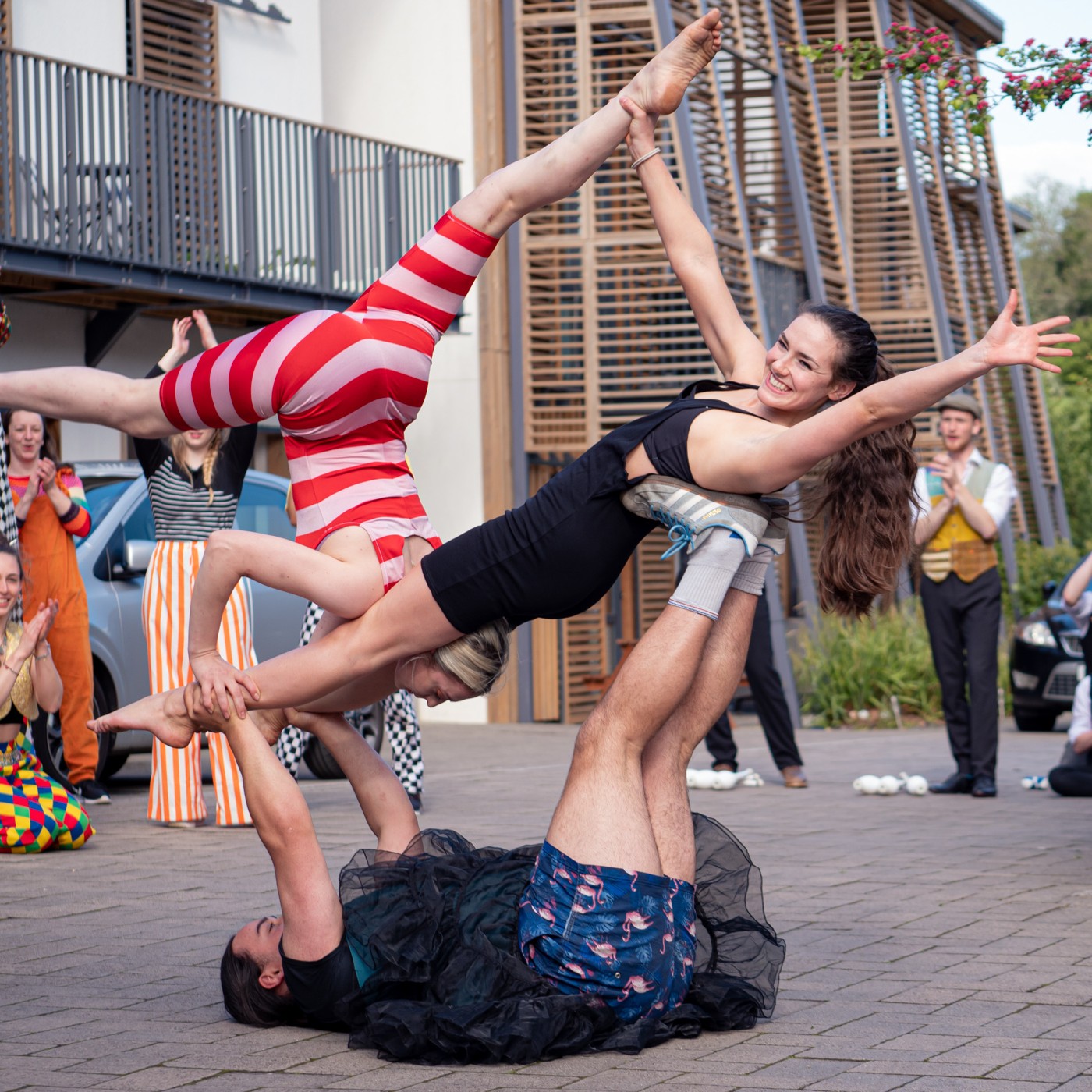 A group of circus performers put on an acrobatic display outdoors. One performer lies on the ground supporting two others balanced mid-air—one in a black outfit and one in red and white stripes. Both are smiling with arms outstretched as a small crowd claps and cheers in the background.